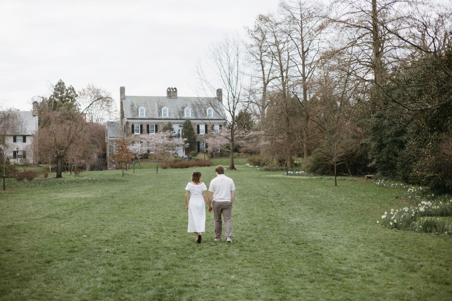Couple walks through a lush green lawn towards a classic white colonial house on a misty spring day.