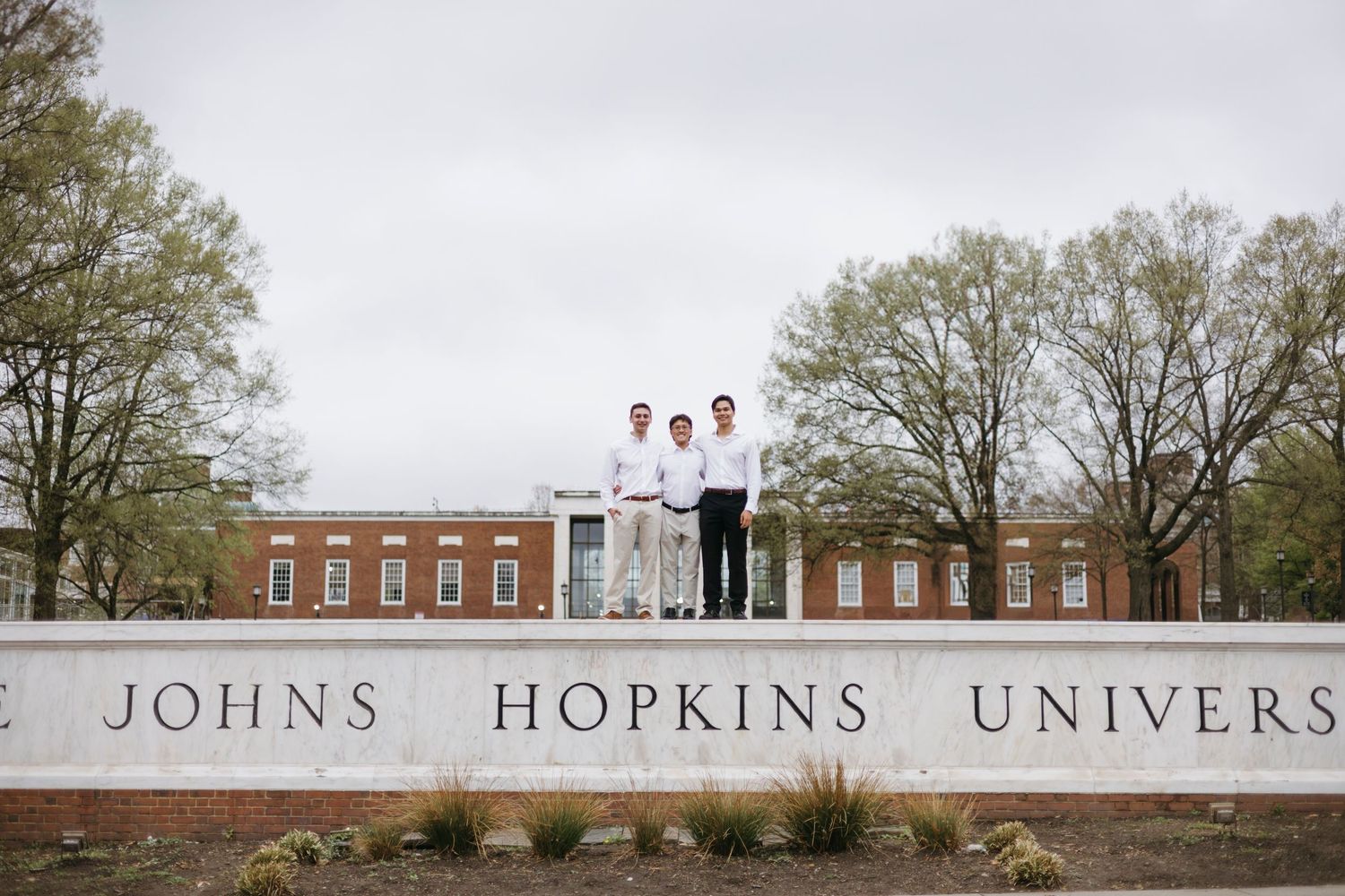 Three people in white lab coats stand on the Johns Hopkins University entrance sign on a cloudy day.