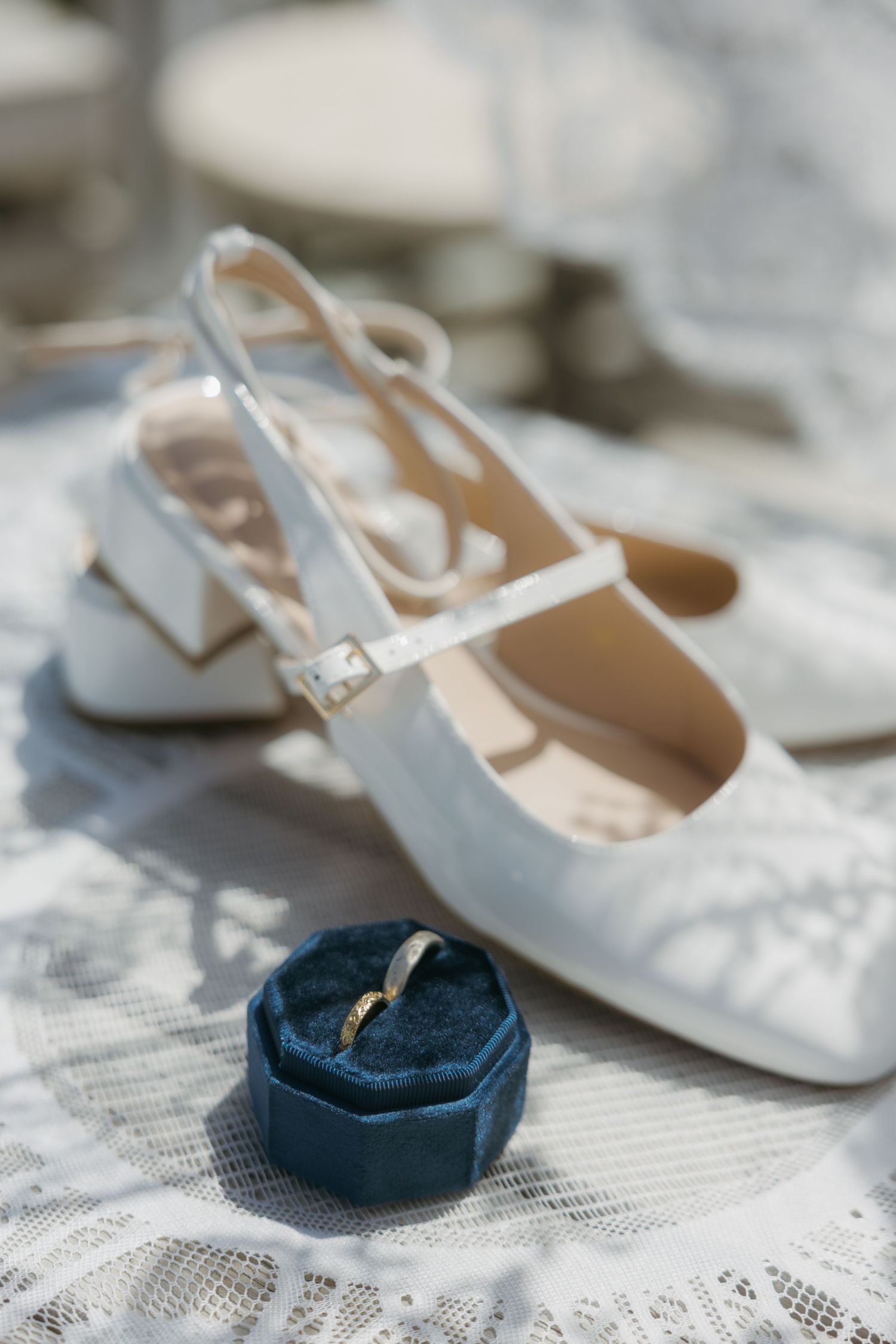 White bridal slingback heels with a gold wedding band in a blue velvet ring box on lace fabric.