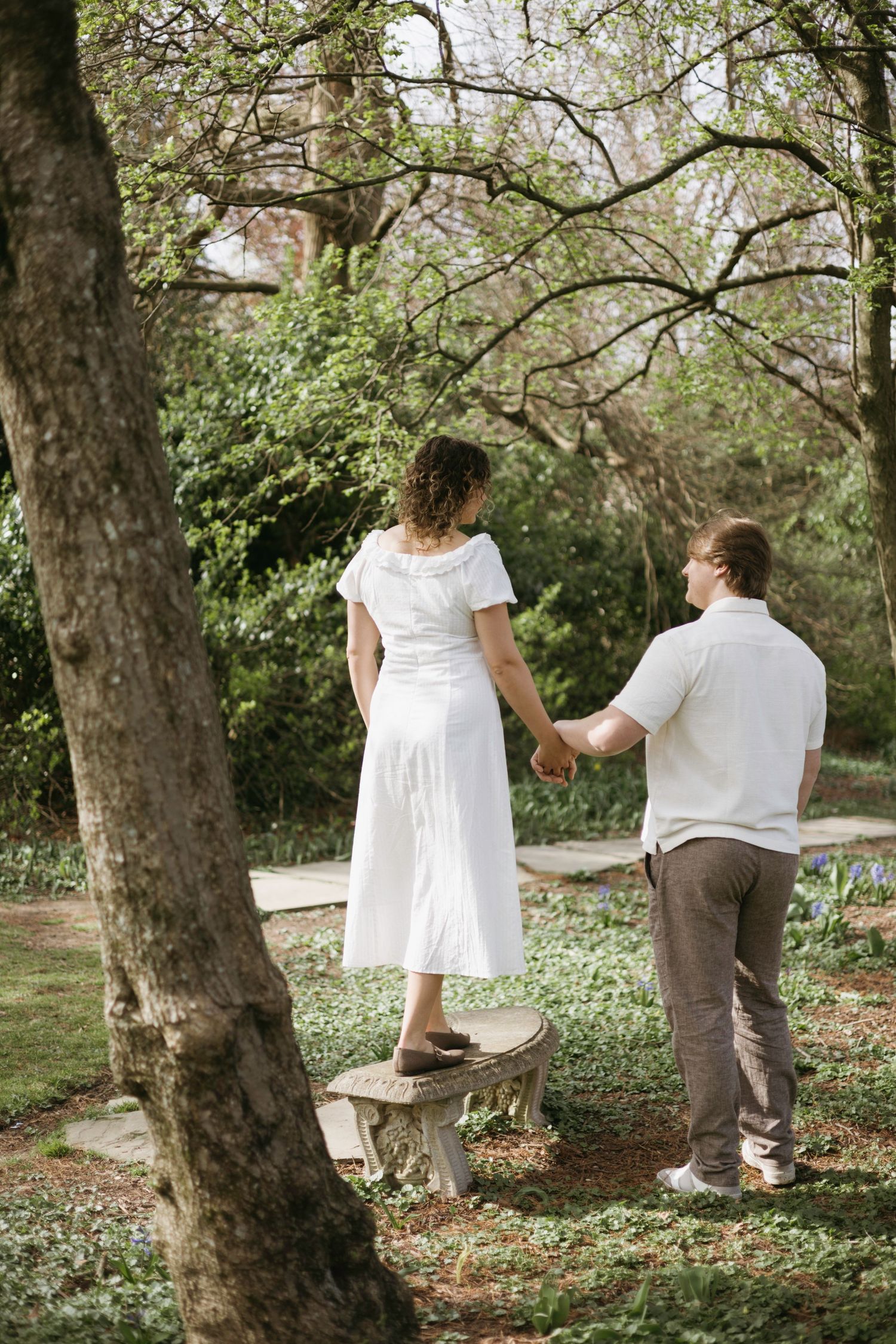 A couple holding hands in a garden setting with tall trees and spring flowers, with one person standing on a stone bench.