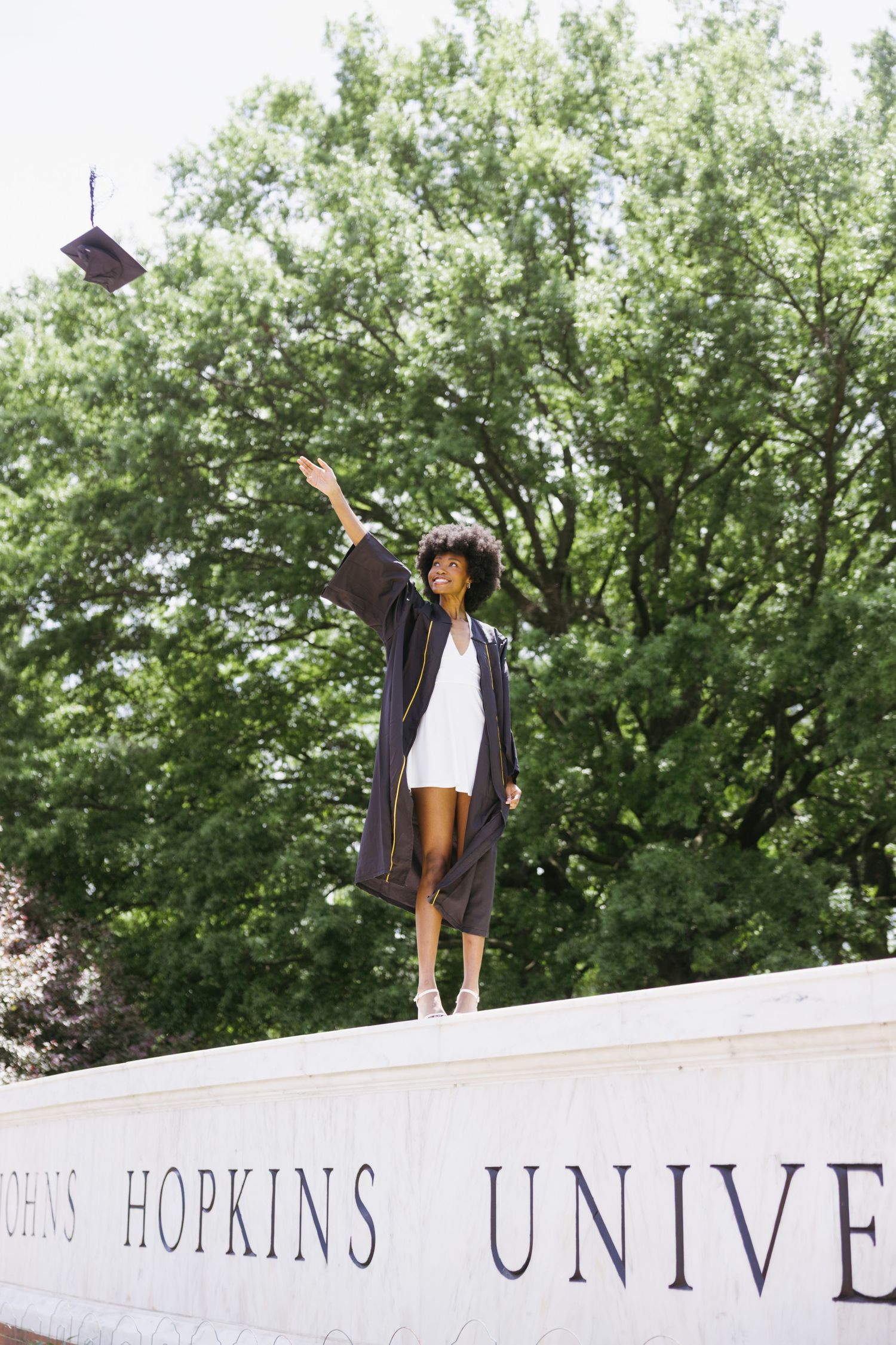 Graduate posing in black cap and gown against Johns Hopkins University sign on sunny day with trees in background.