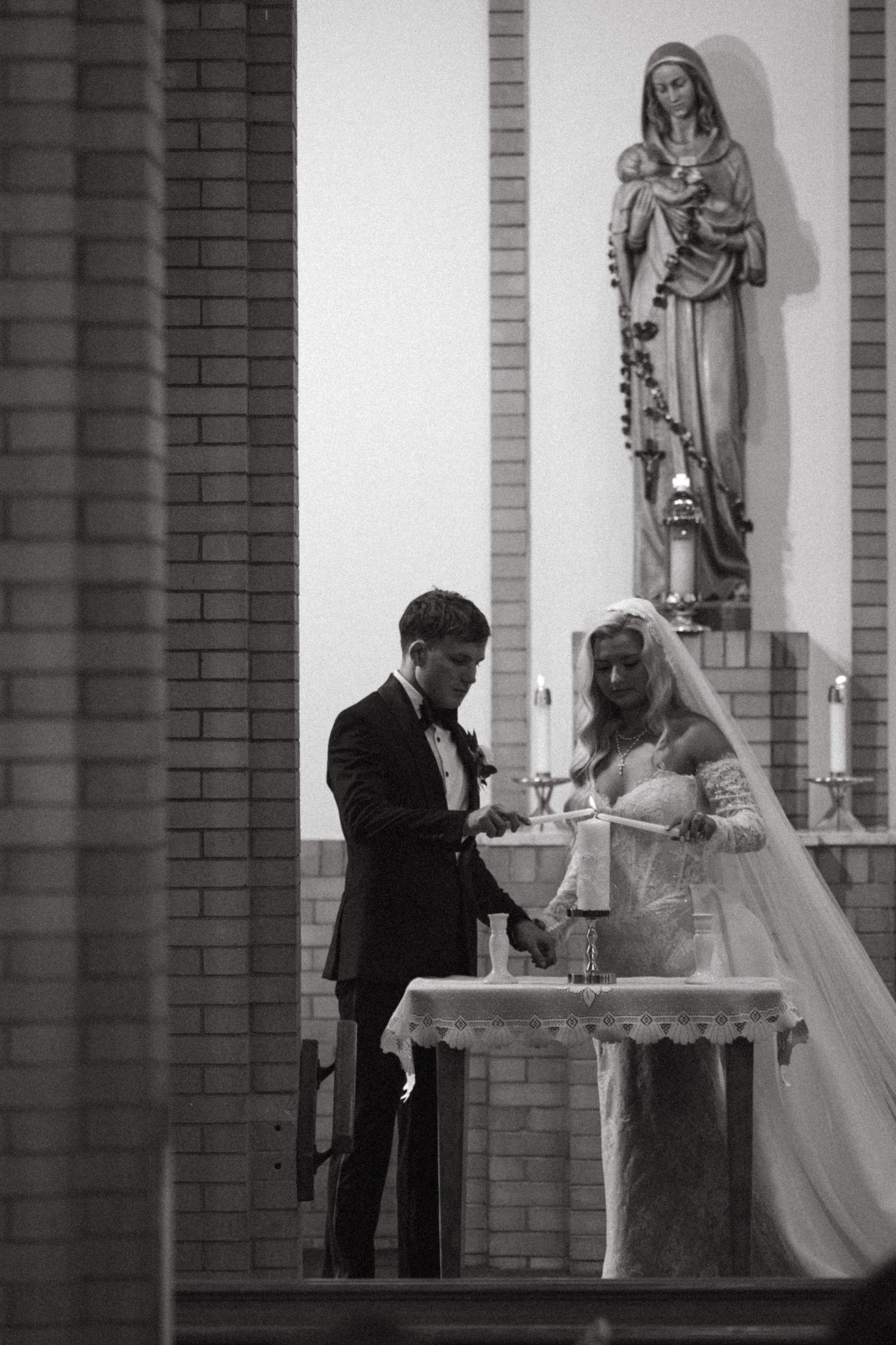 Black and white photo of a bride and groom lighting a unity candle at a Catholic church altar during their wedding ceremony.