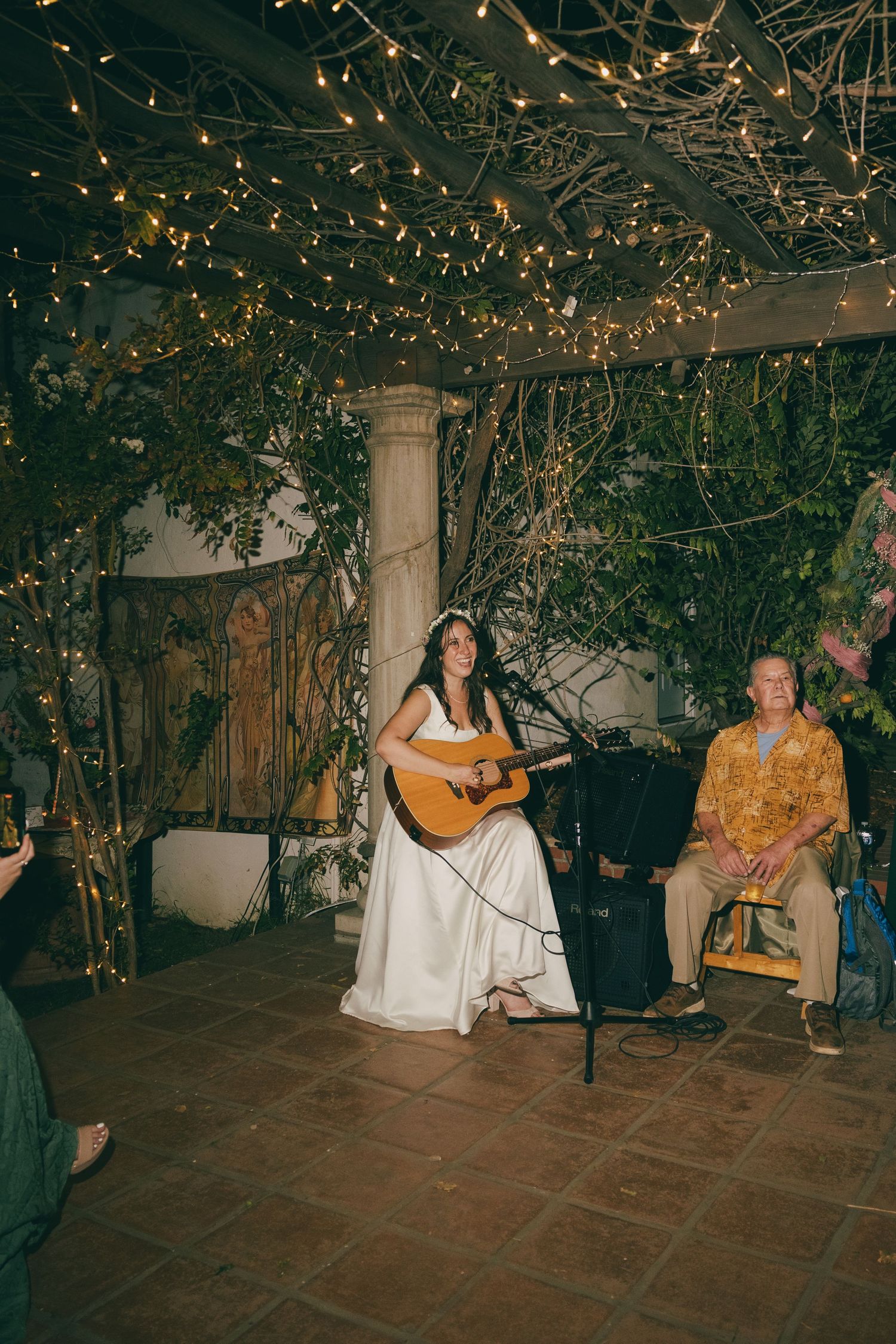 Bride in white gown playing acoustic guitar at outdoor wedding reception with fairy lights and greenery backdrop.