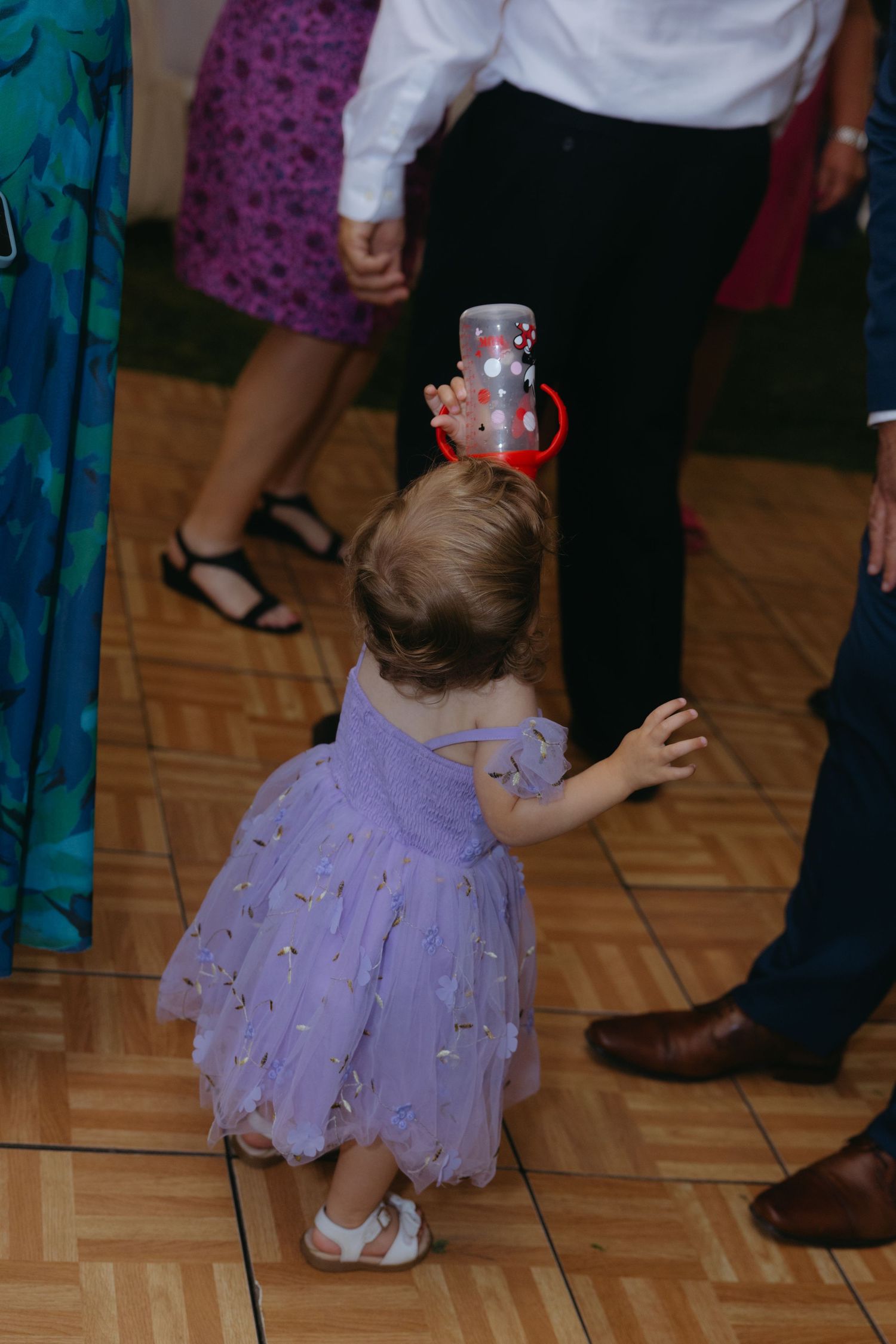 A small child in a purple tulle dress crouches down on a hardwood dance floor during a celebration.