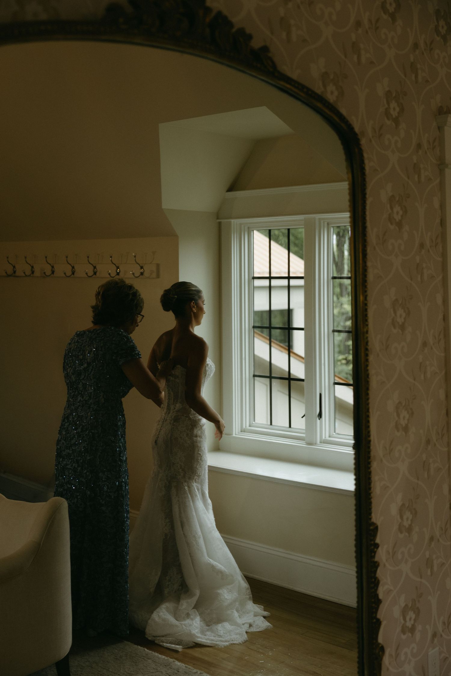 Bride in white lace gown stands by window as woman helps with dress, reflected in ornate gold mirror.