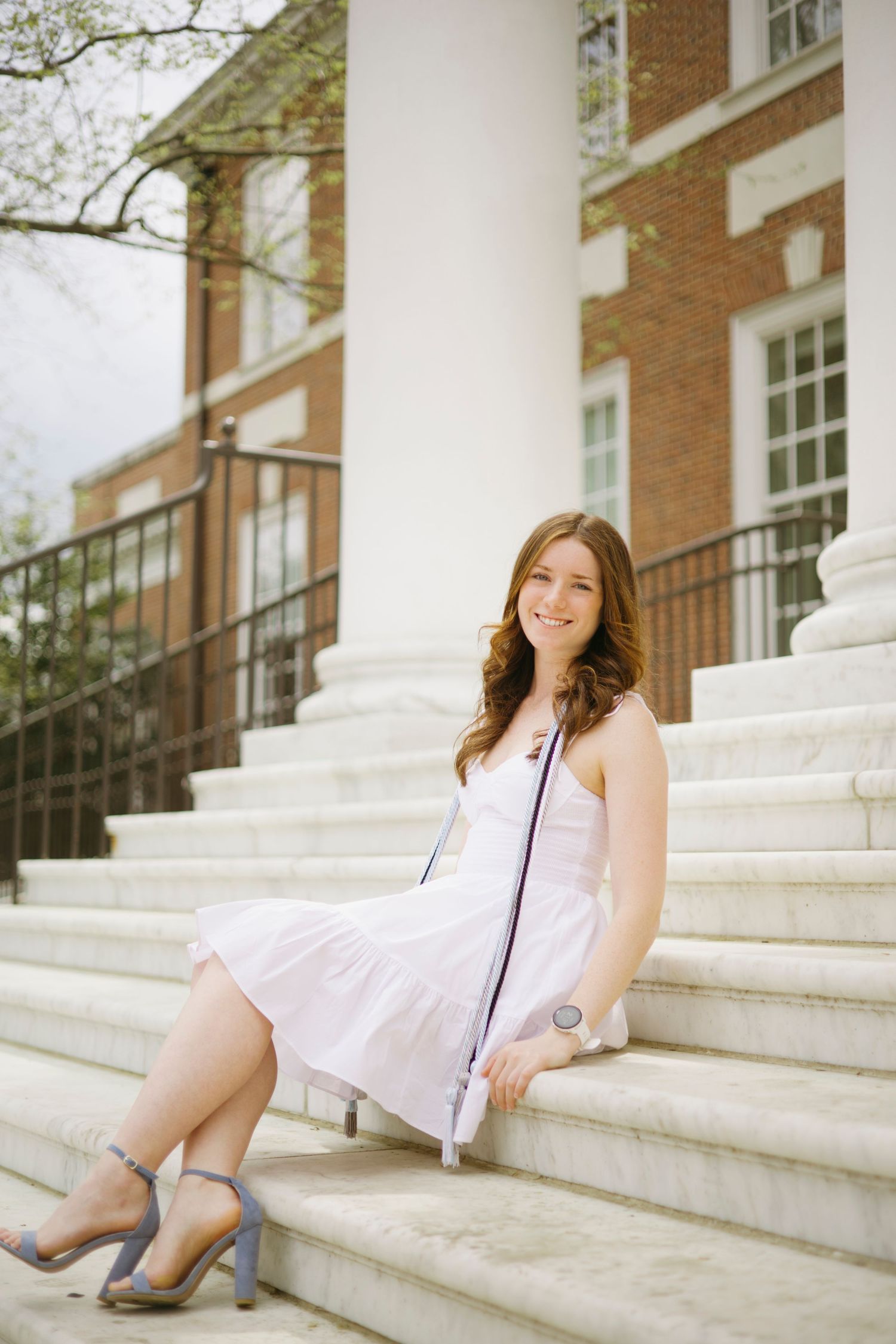 A person in a white dress sitting on outdoor stone steps with columns and brick building in background.