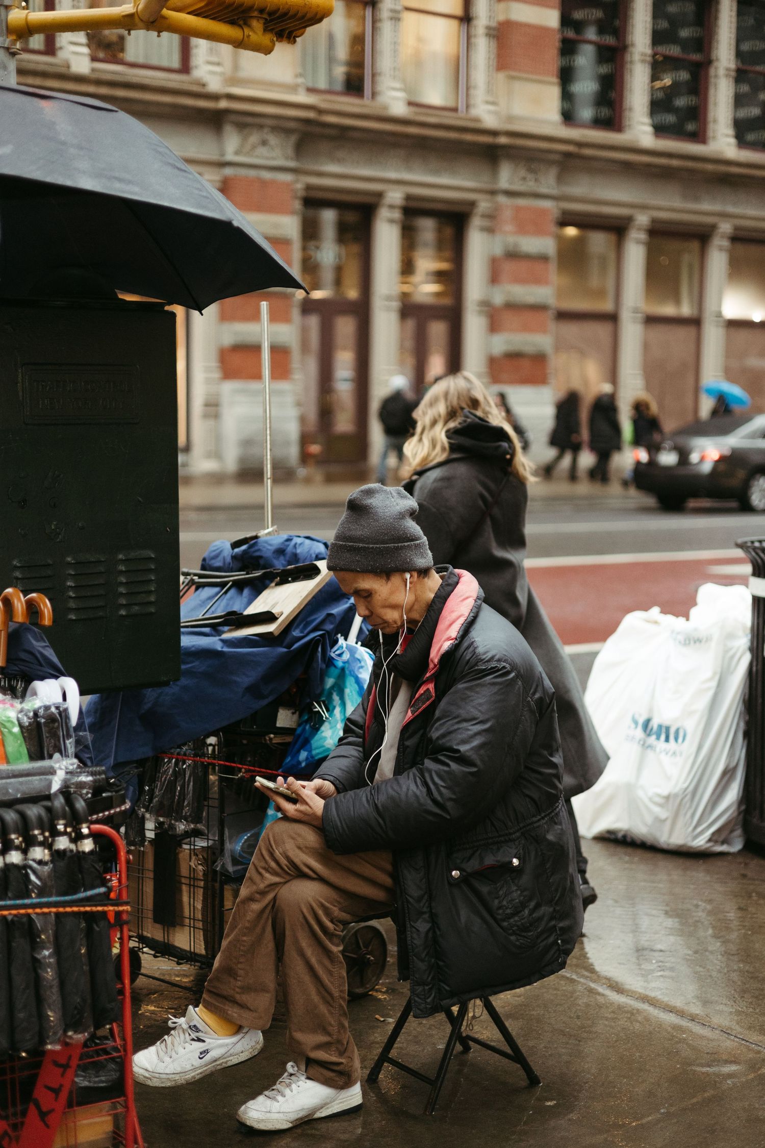 Street vendor sitting on a folding chair checks phone while managing their outdoor stand on a rainy city sidewalk.