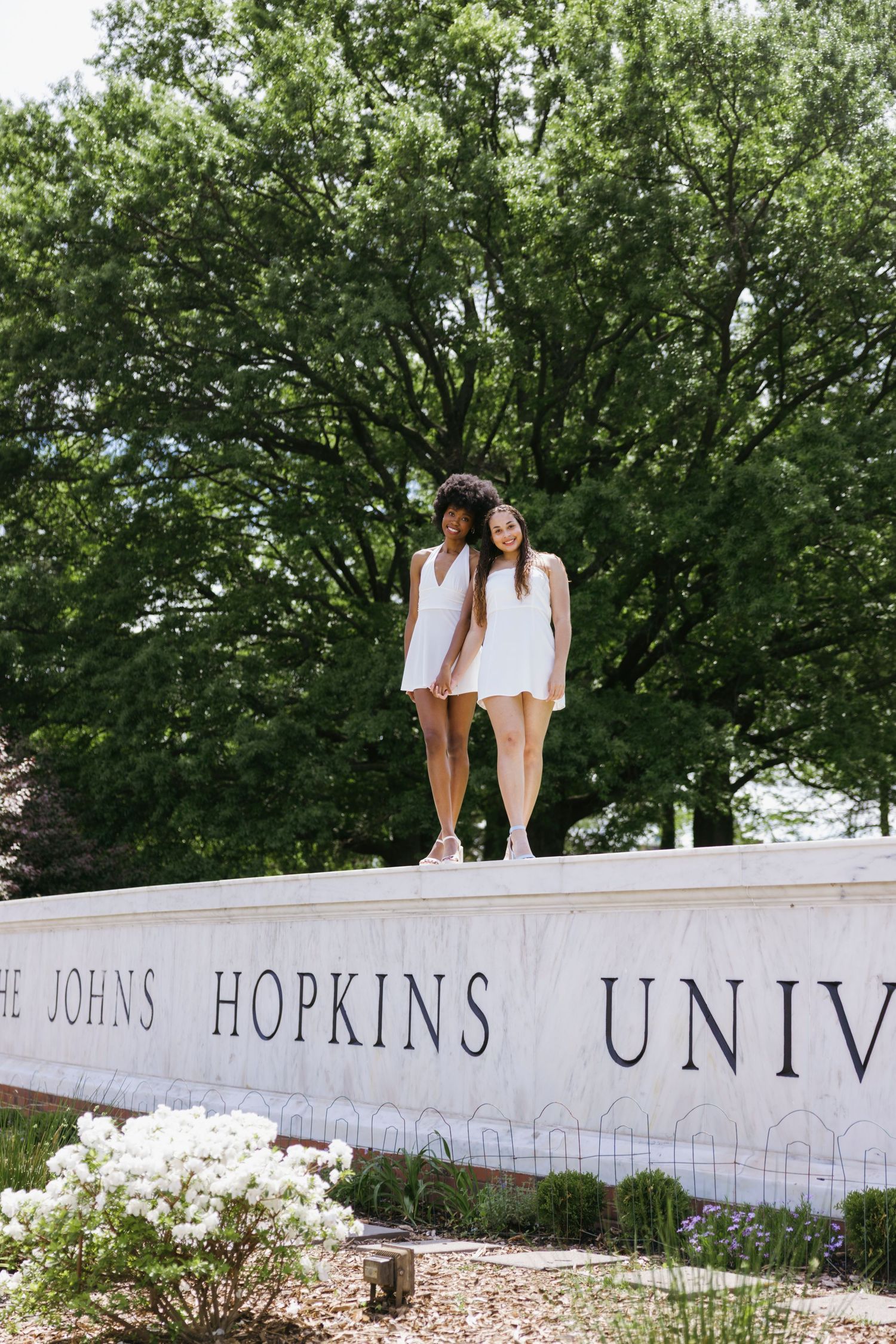 Two students in white outfits stand on Johns Hopkins University entrance sign against lush green trees in background.