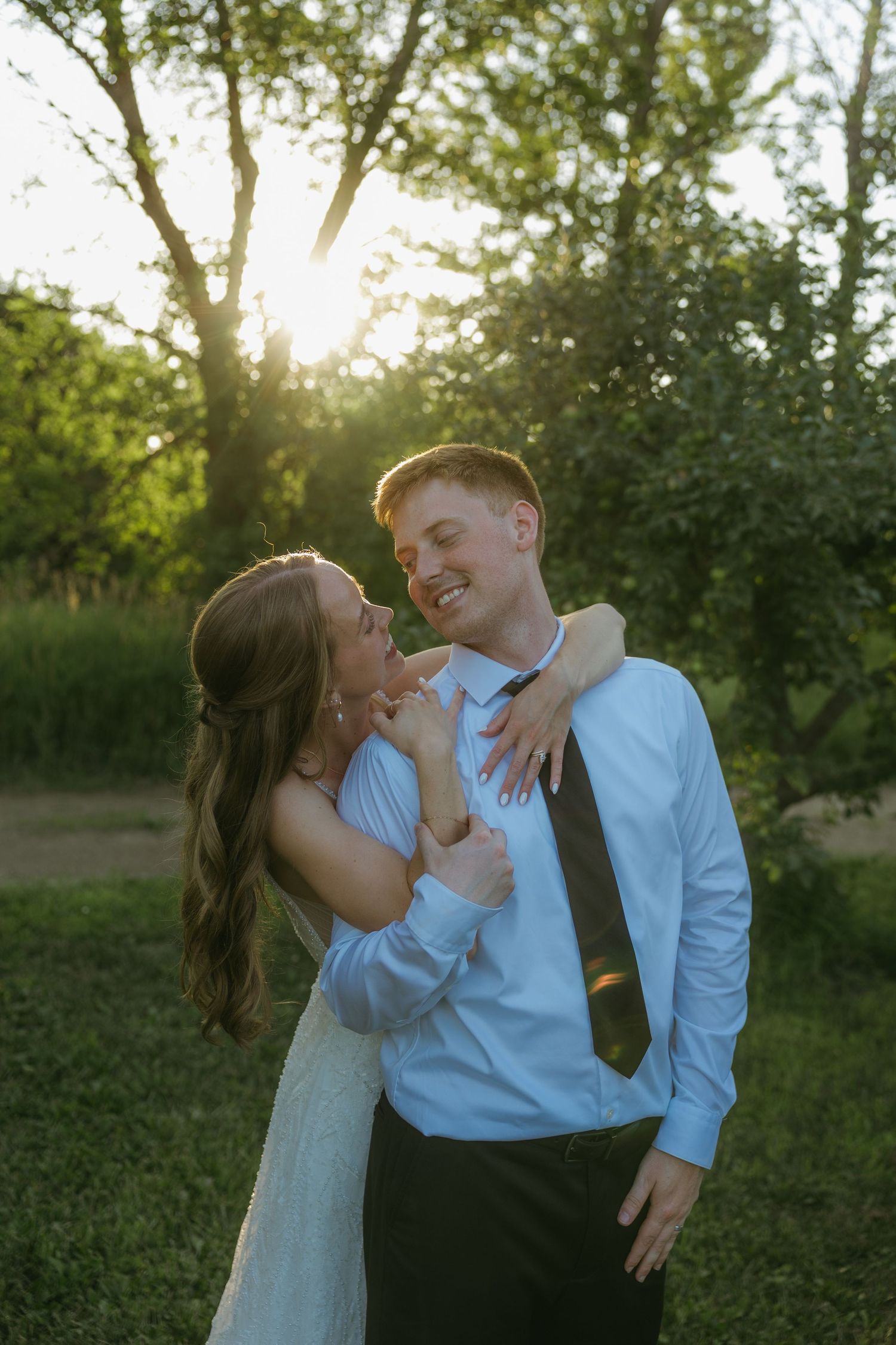 A couple shares a romantic moment outdoors at golden hour, the groom in a white shirt and tie smiling as the bride embraces him.