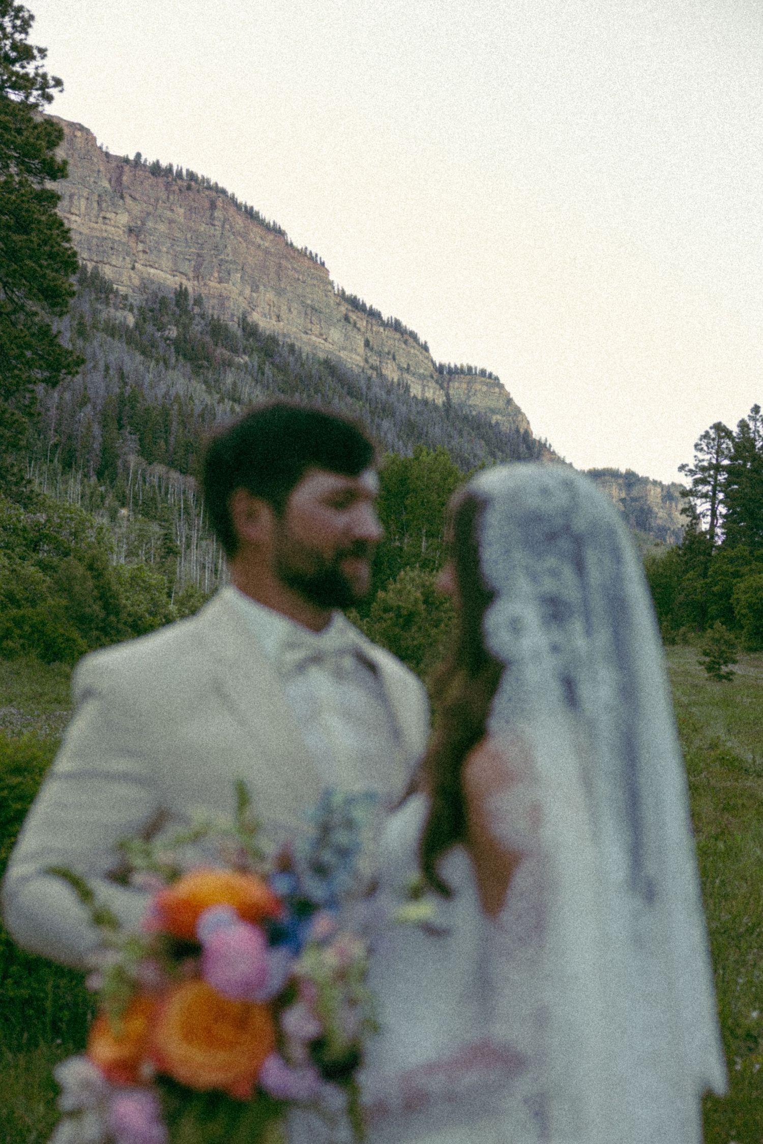 Bride and groom in mountain meadow, groom in white suit, bride holding colorful bouquet with rocky cliffs backdrop.