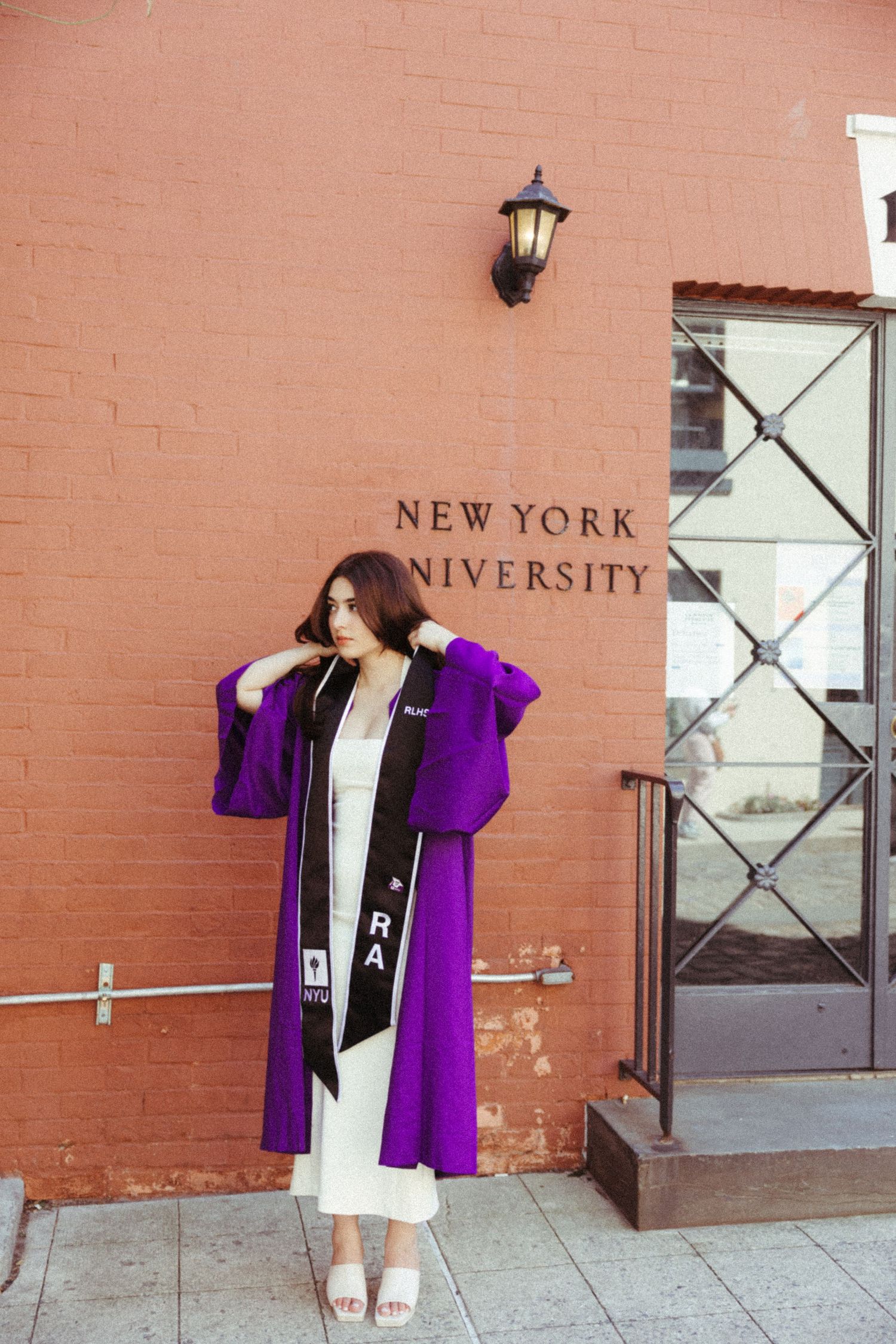 Graduate in a purple academic regalia posing against NYU brick wall with vintage lamp.