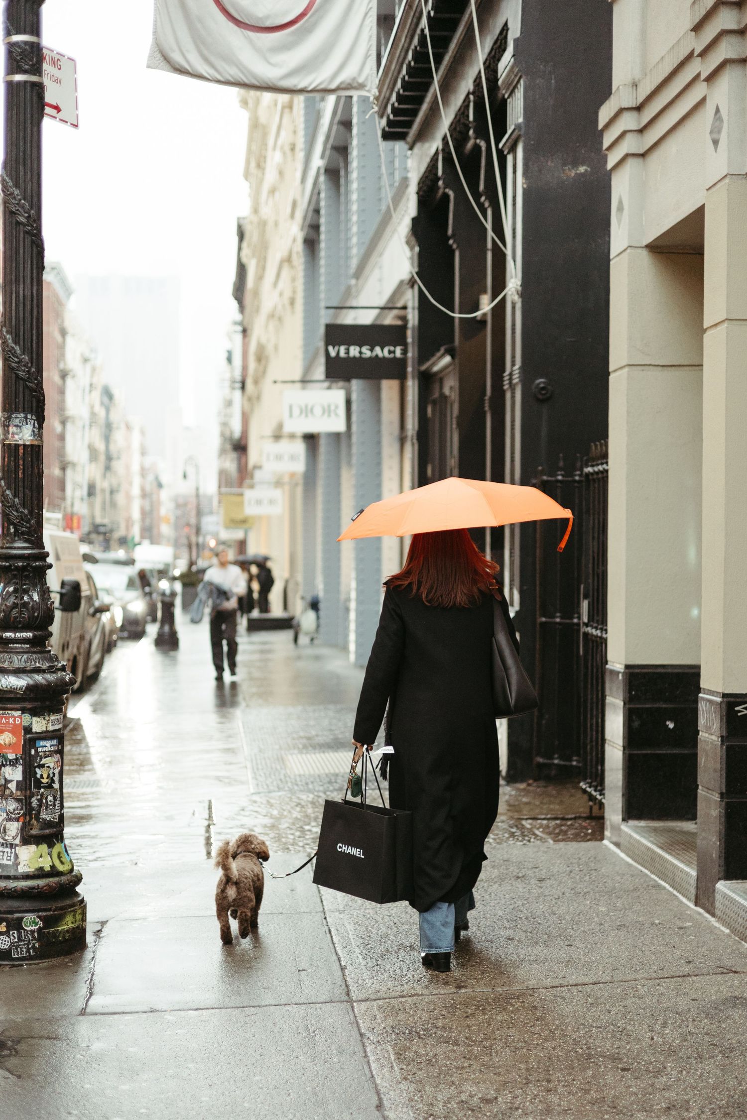 Someone in a long black coat walks their small brown dog on a rainy city street while holding an orange umbrella.