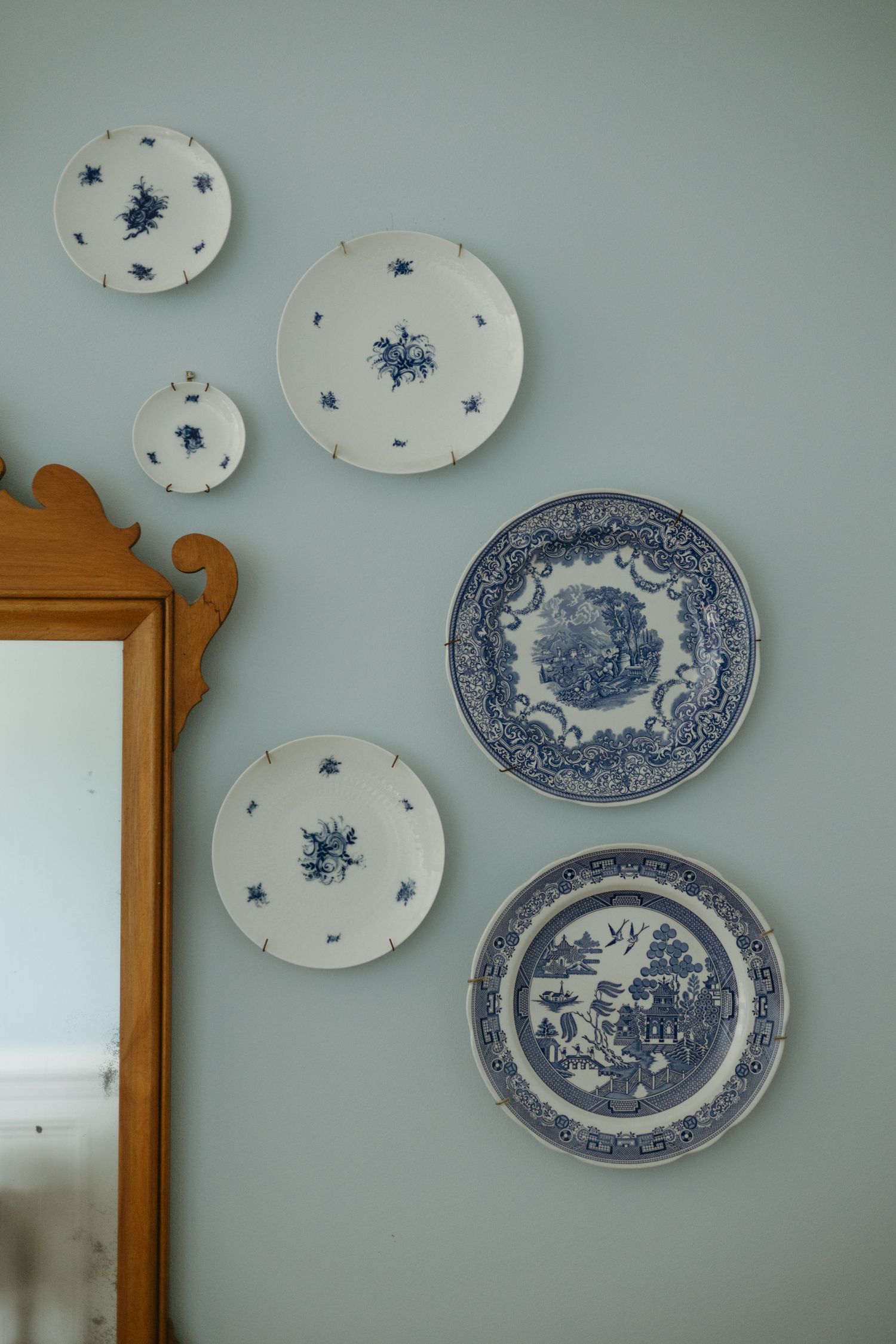 Decorative blue and white antique plates arranged on a light blue wall next to a wooden framed mirror.