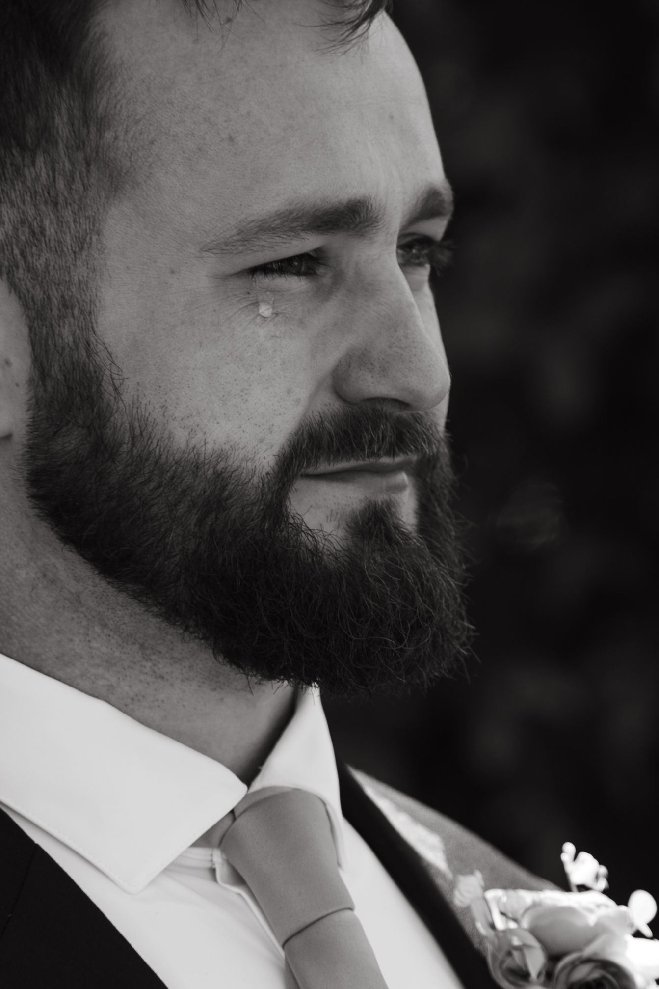 Black and white portrait of a bearded groom crying with a tear on his cheek, wearing a suit and floral boutonniere.