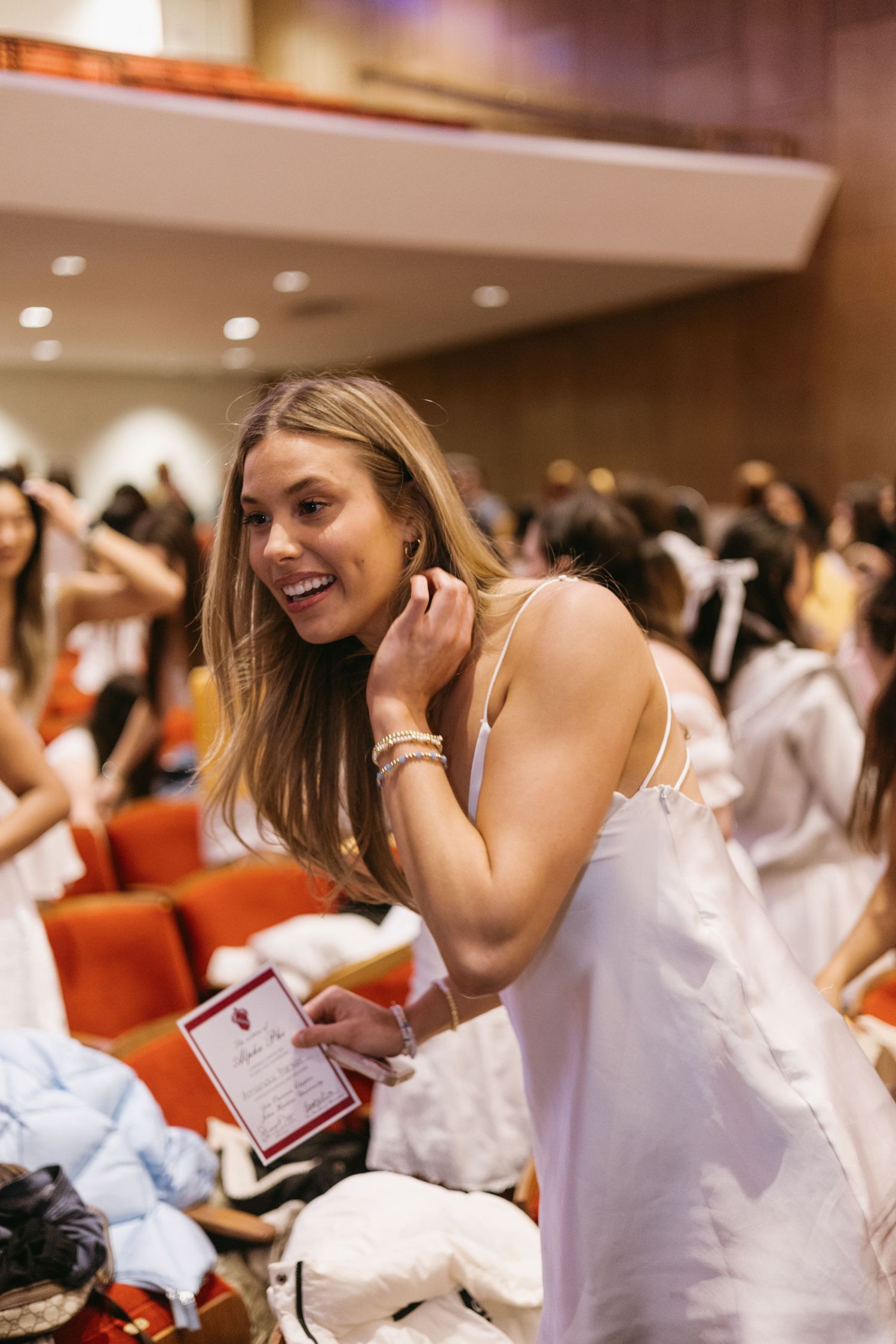A person in a white dress smiles while seated in an auditorium among a crowd of other attendees.