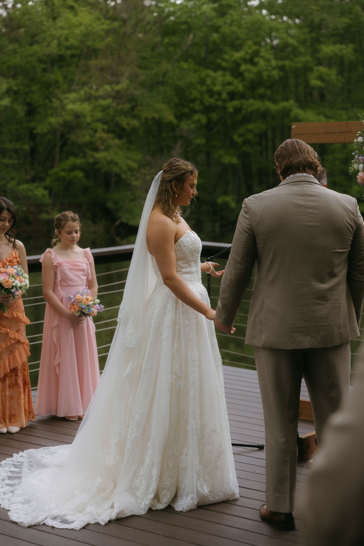 Bride in lace gown holds grooms hands on outdoor deck during wedding ceremony with bridesmaids in pink and orange dresses.
