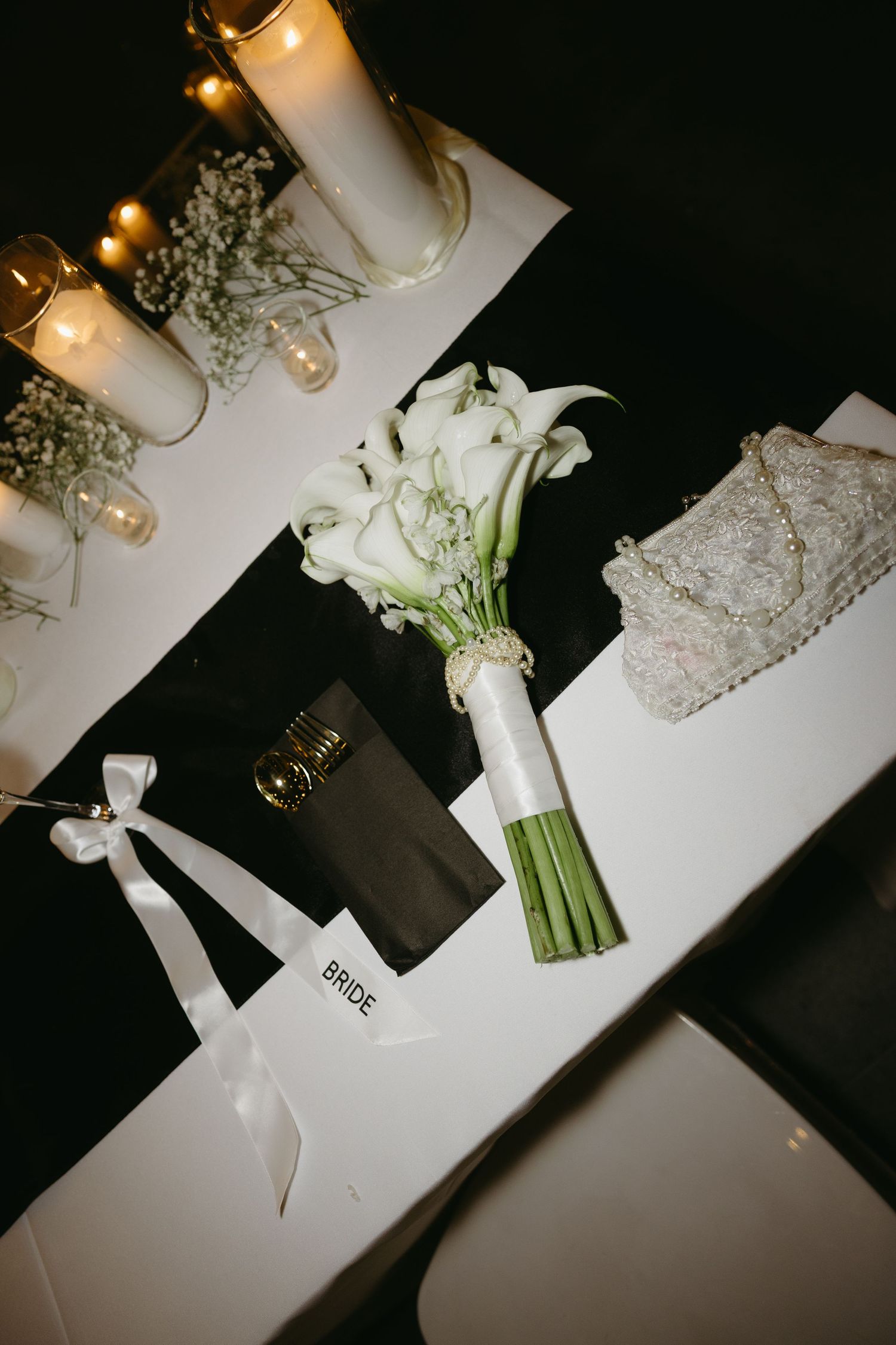 Elegant wedding flat lay with white calla lily bridal bouquet, candles, gold cutlery, and a bride tote bag on a table.