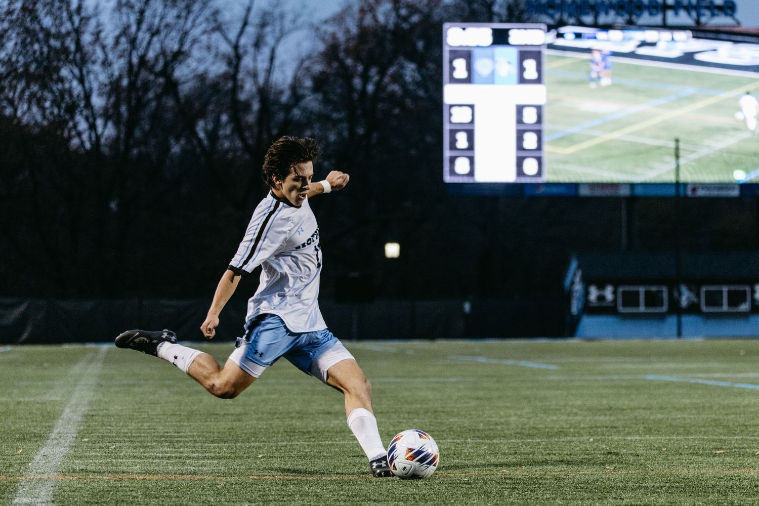 Soccer player kicks ball during evening match with scoreboard visible in background.