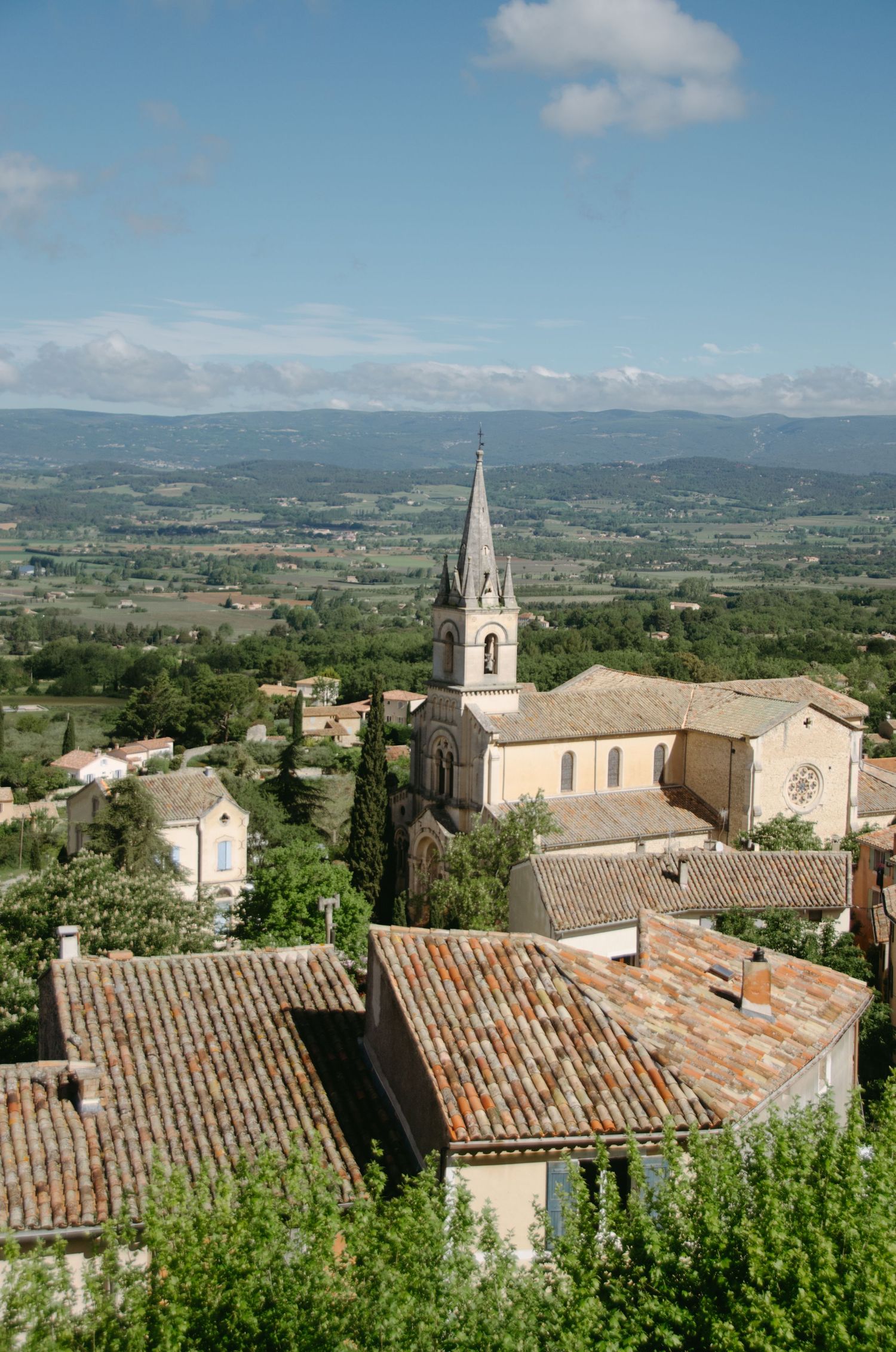 Church spire rises above terracotta roofs in French countryside with mountains in background.