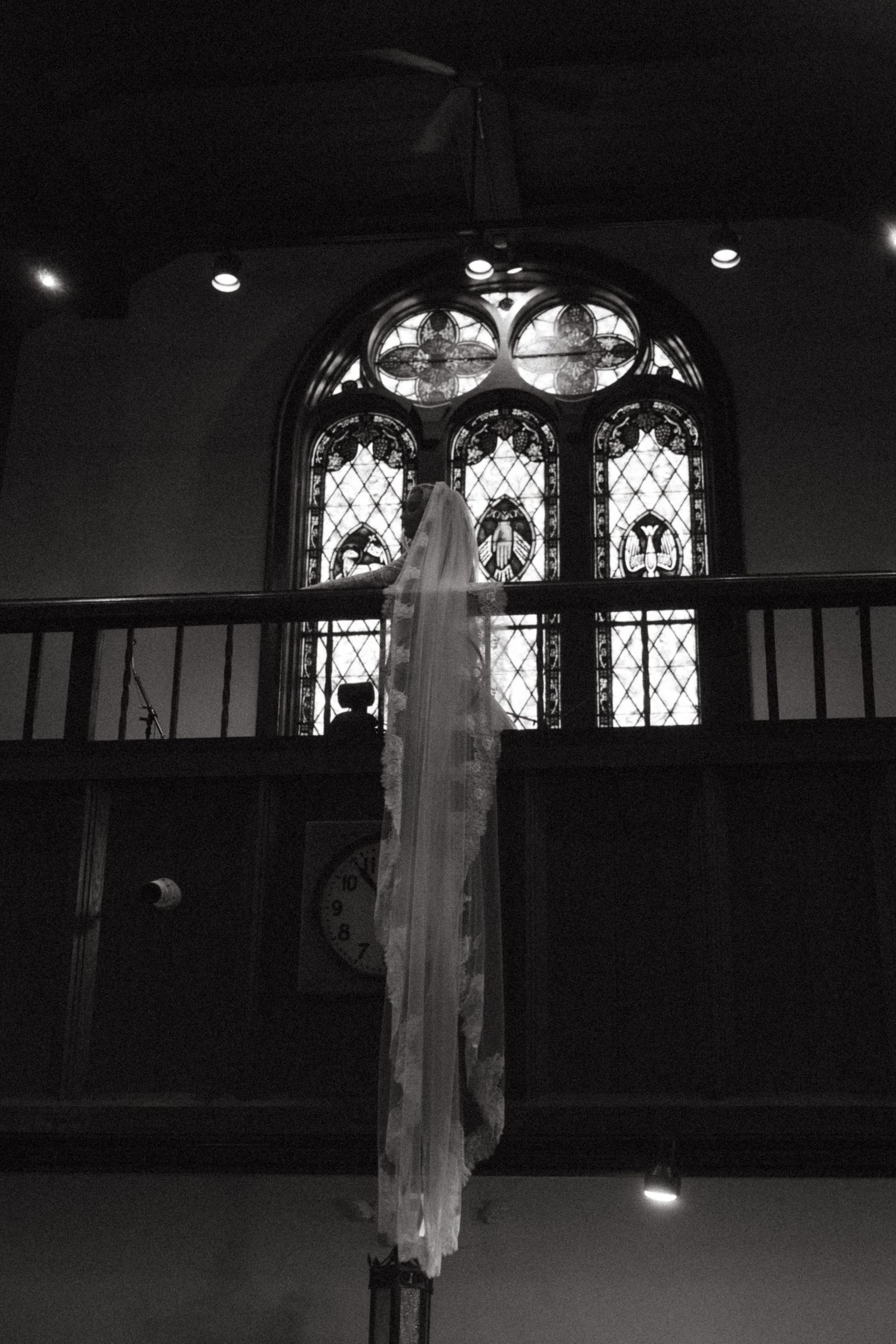 Black and white photo of a bridal veil hanging in a church with stained glass windows and arched architecture.