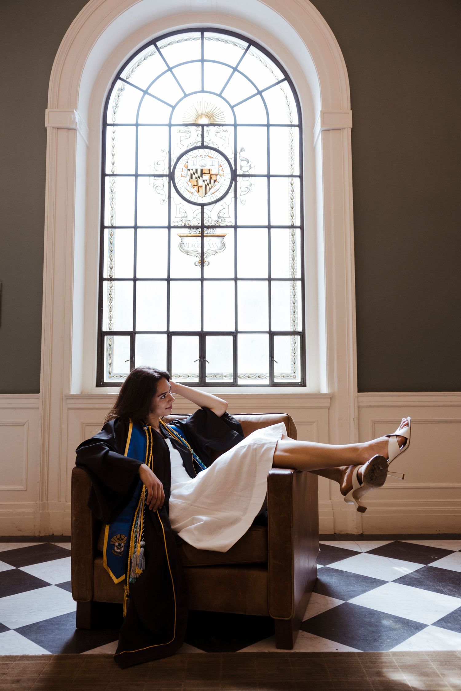 Graduate in cap and gown relaxes in vintage chair beneath ornate arched window with checkerboard floor.