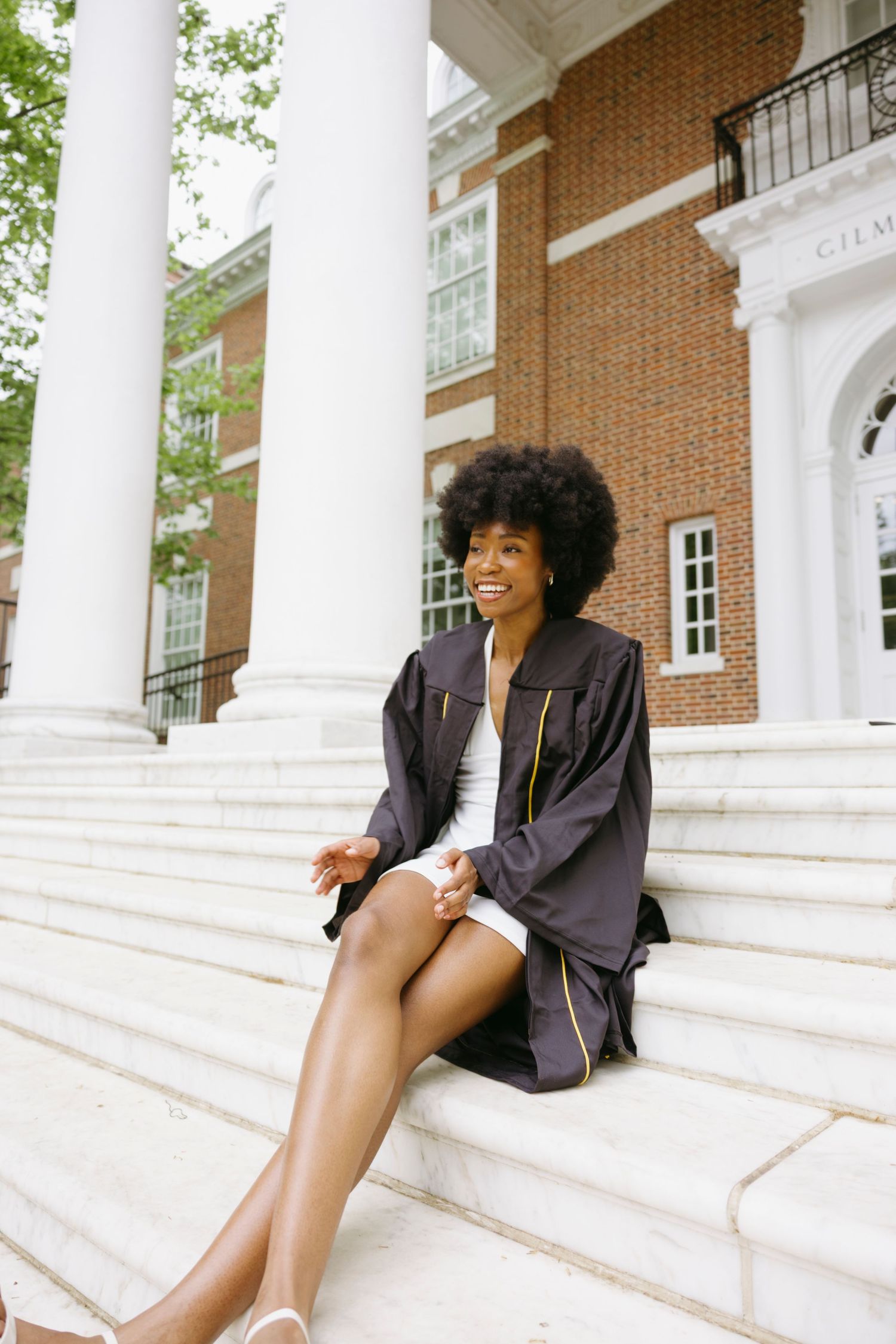 A graduate in a black gown sits smiling on white marble steps in front of a classic brick university building with columns.