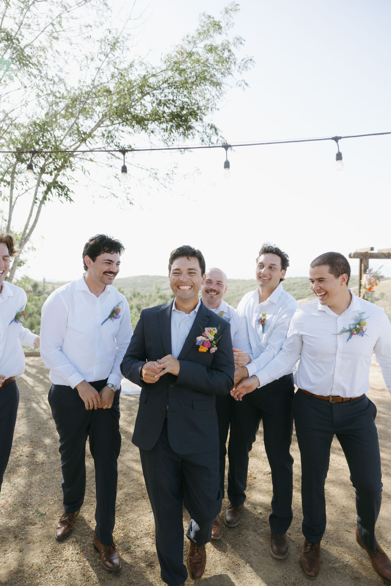 Groom in navy suit walks with smiling groomsmen in white shirts outdoors at a sunny wedding venue.