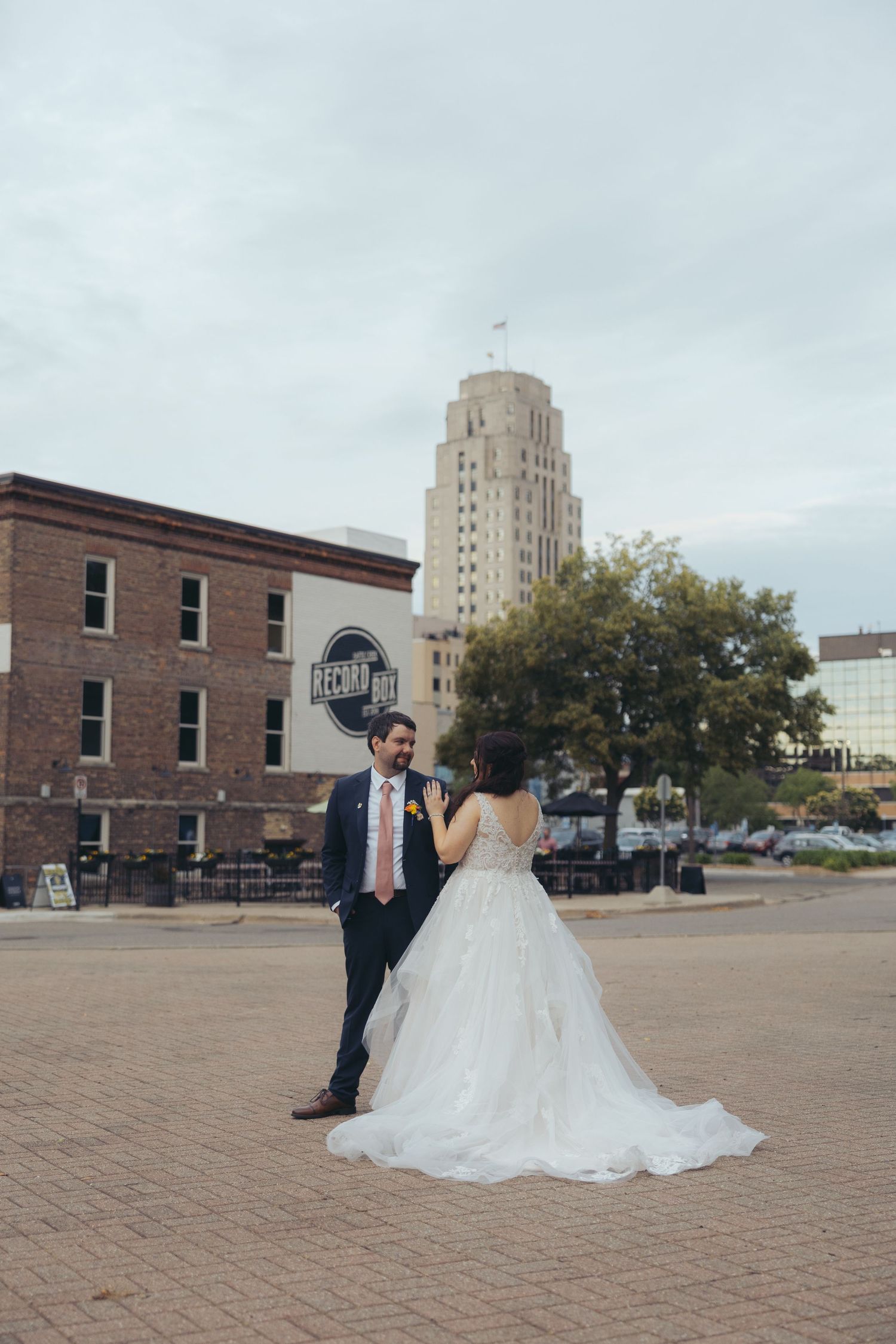 Bride and groom pose in urban plaza with historic brick building and art deco skyscraper backdrop on their wedding day.