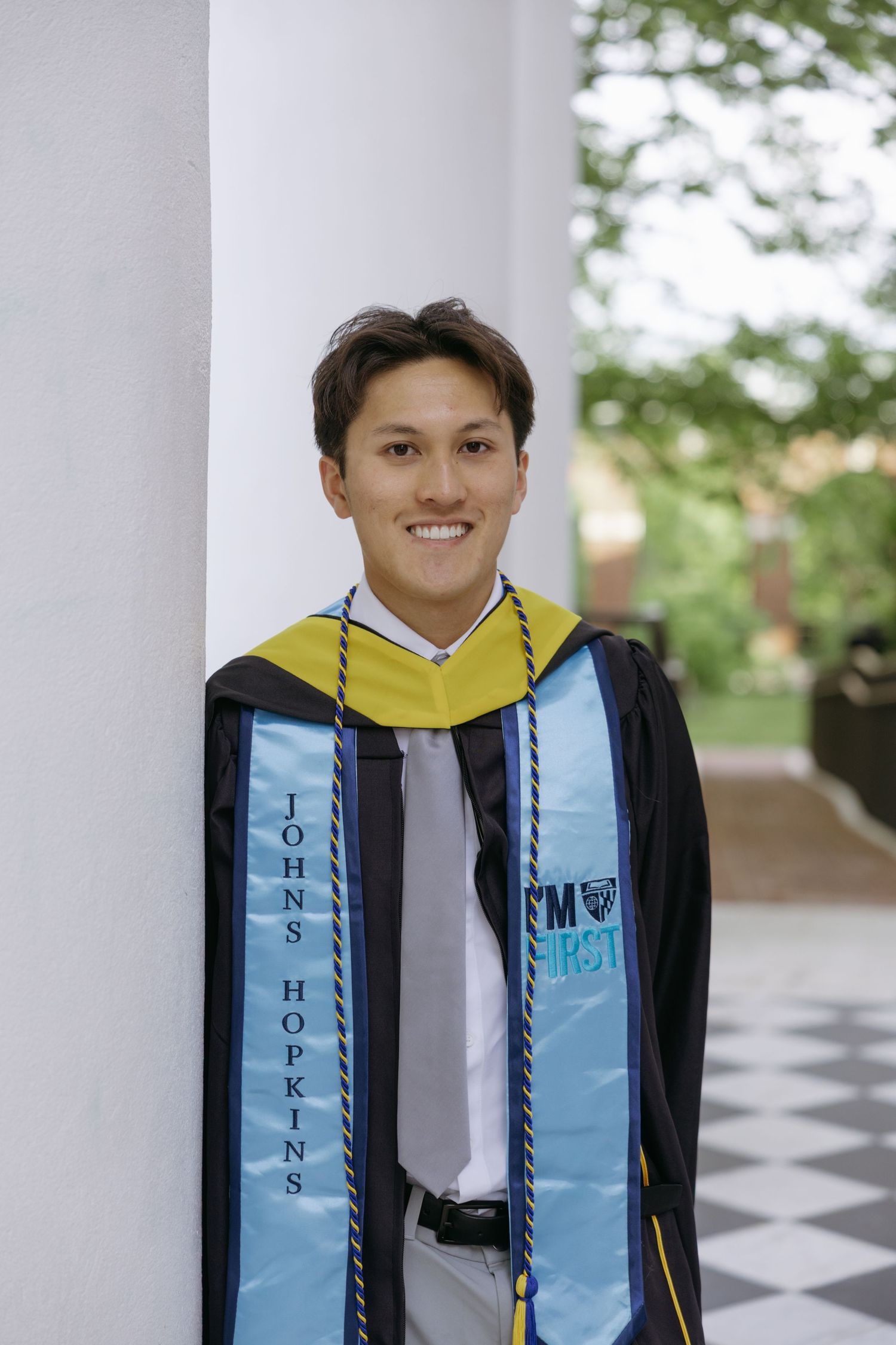Graduate in academic regalia wearing blue sash with Johns Hopkins logo at outdoor ceremony location.