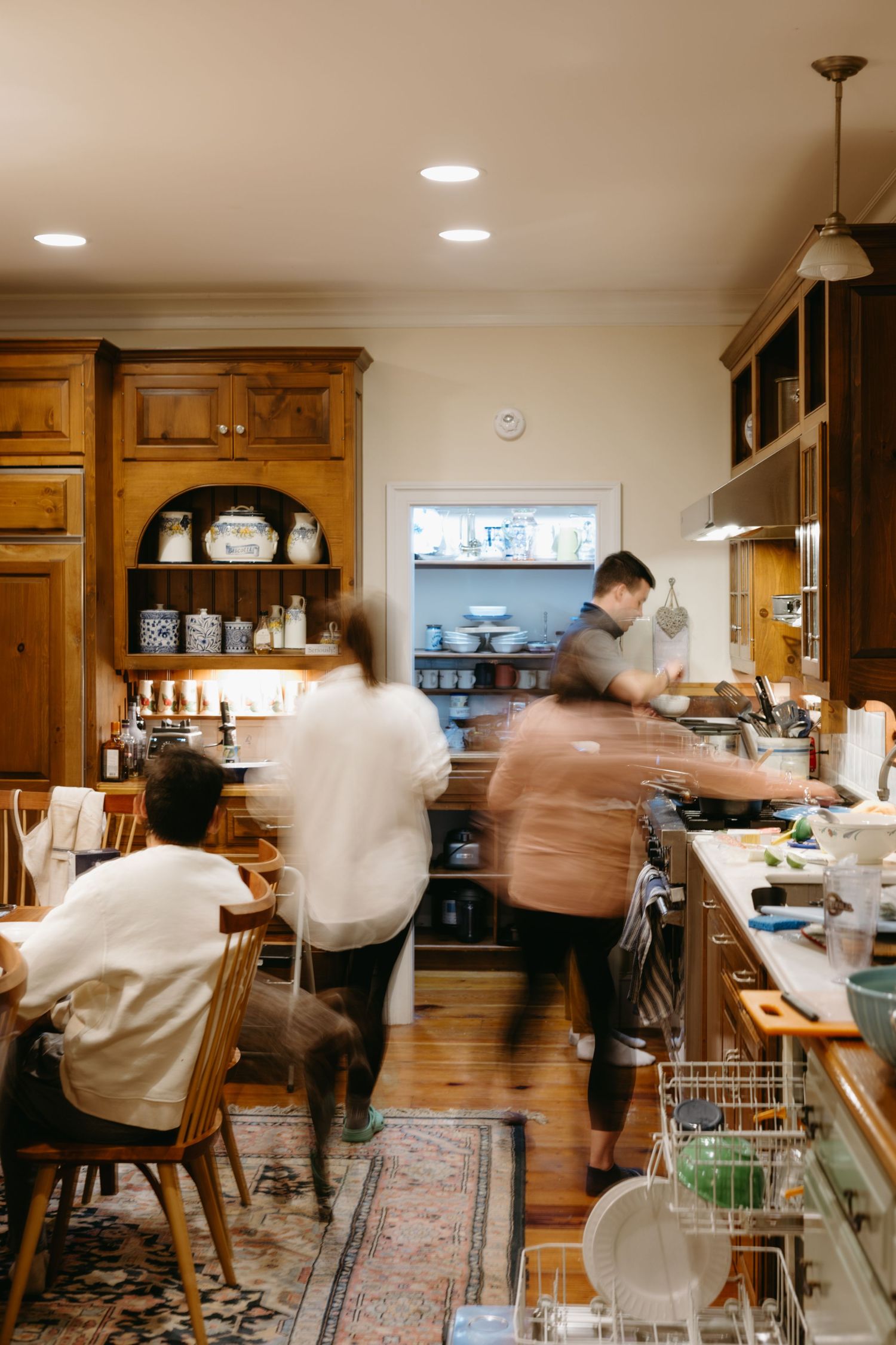 Blurred motion of people working in a cozy kitchen with wooden cabinets, recessed lighting, and vintage style decor on an area rug.