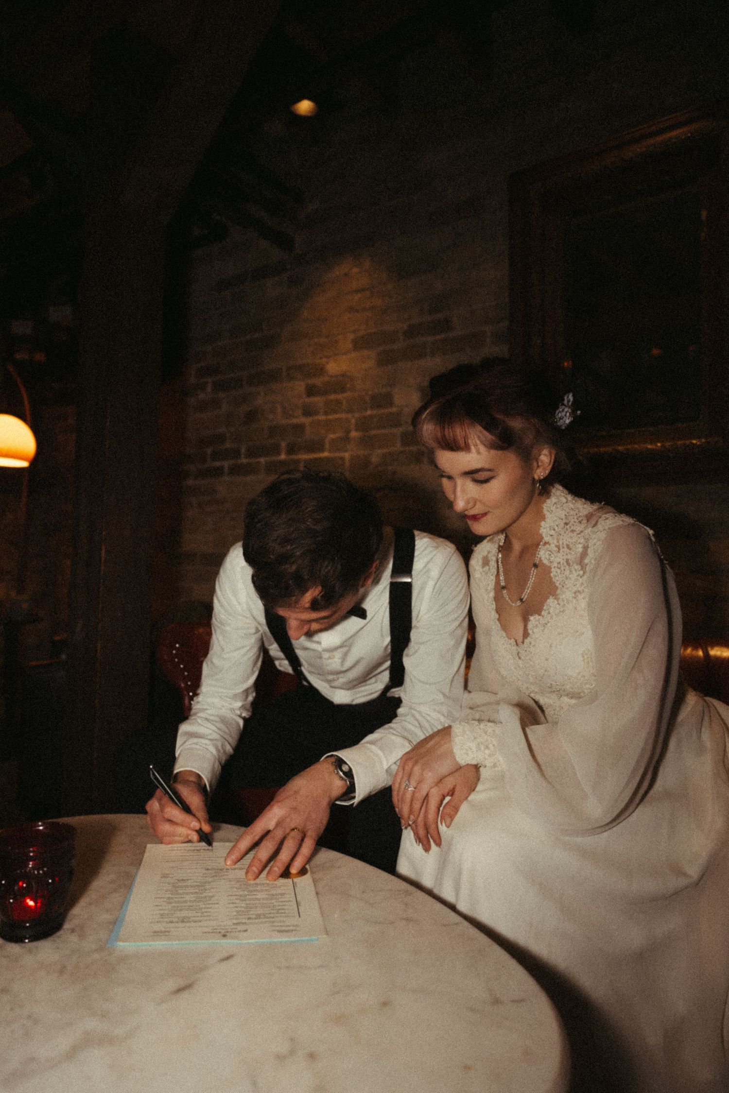 Two people in white clothing lean over a document at a rustic table in a dimly lit brick room.