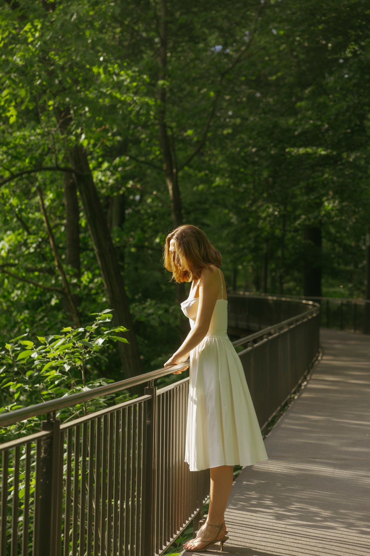 A person in a white summer dress stands contemplatively on a wooden boardwalk surrounded by lush green forest canopy.