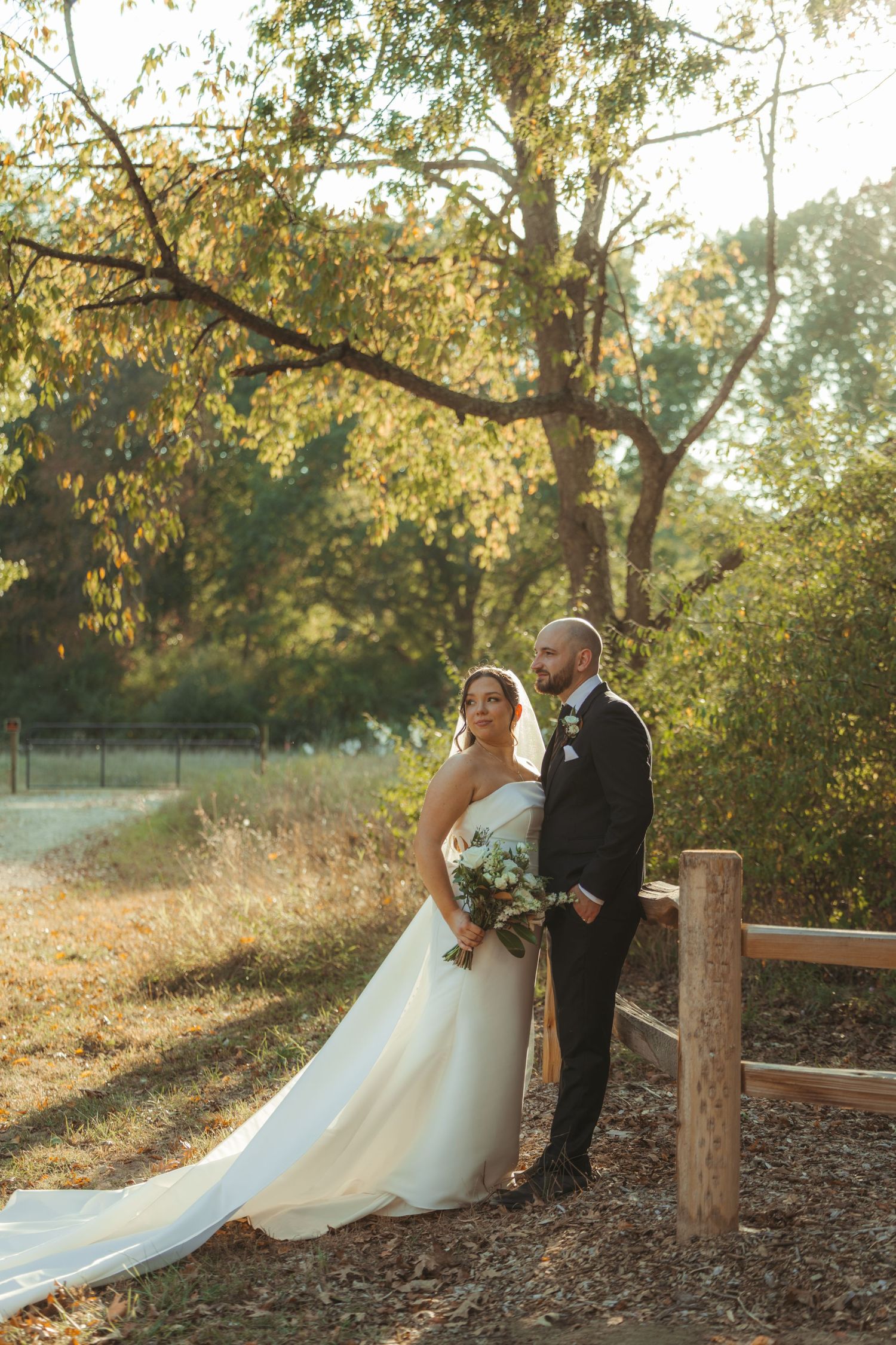A couple in wedding attire poses together at sunset in a rustic outdoor setting with autumn trees and wooden fence posts.