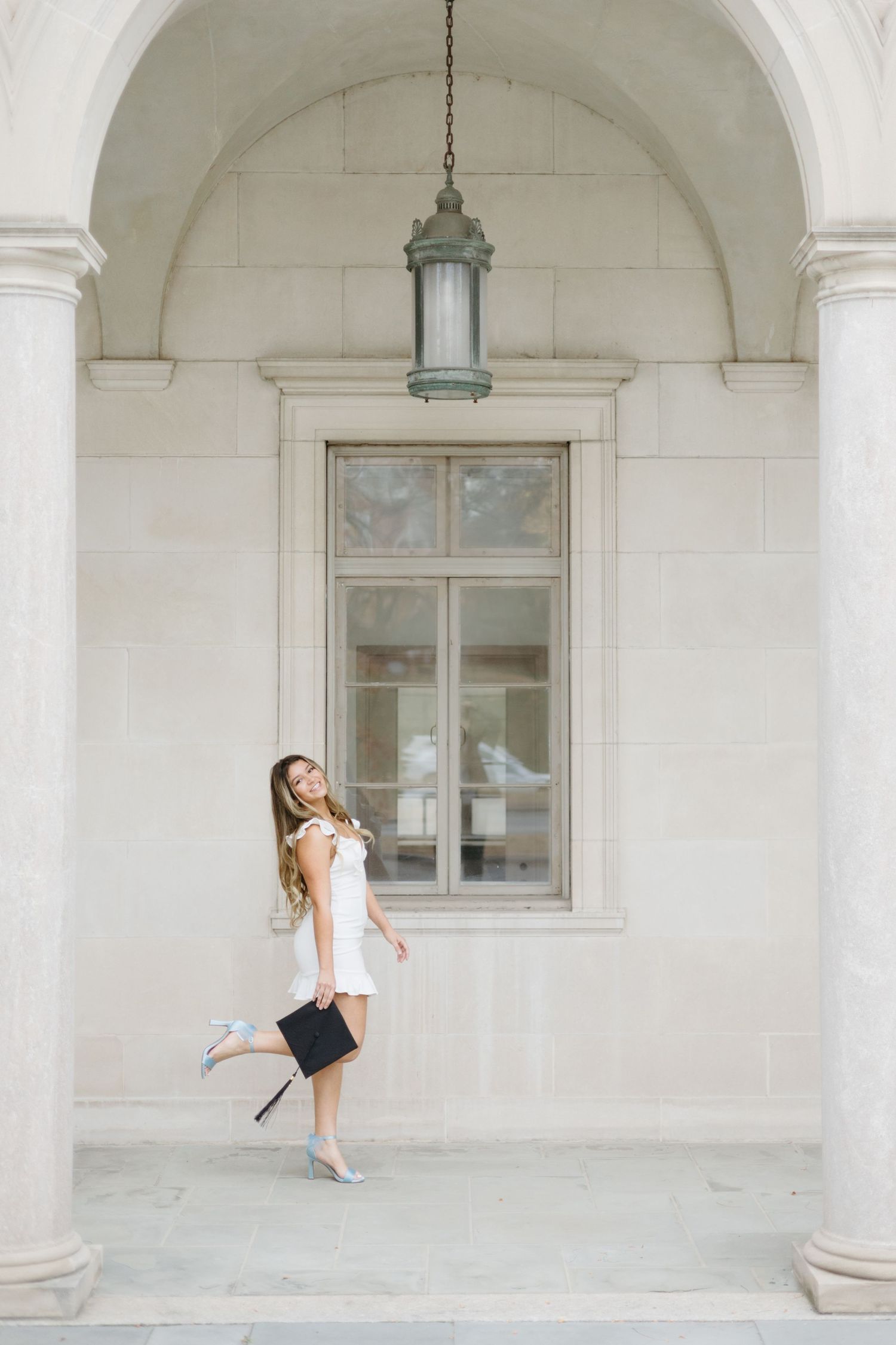 Graduate in white dress poses playfully under arched university entrance.