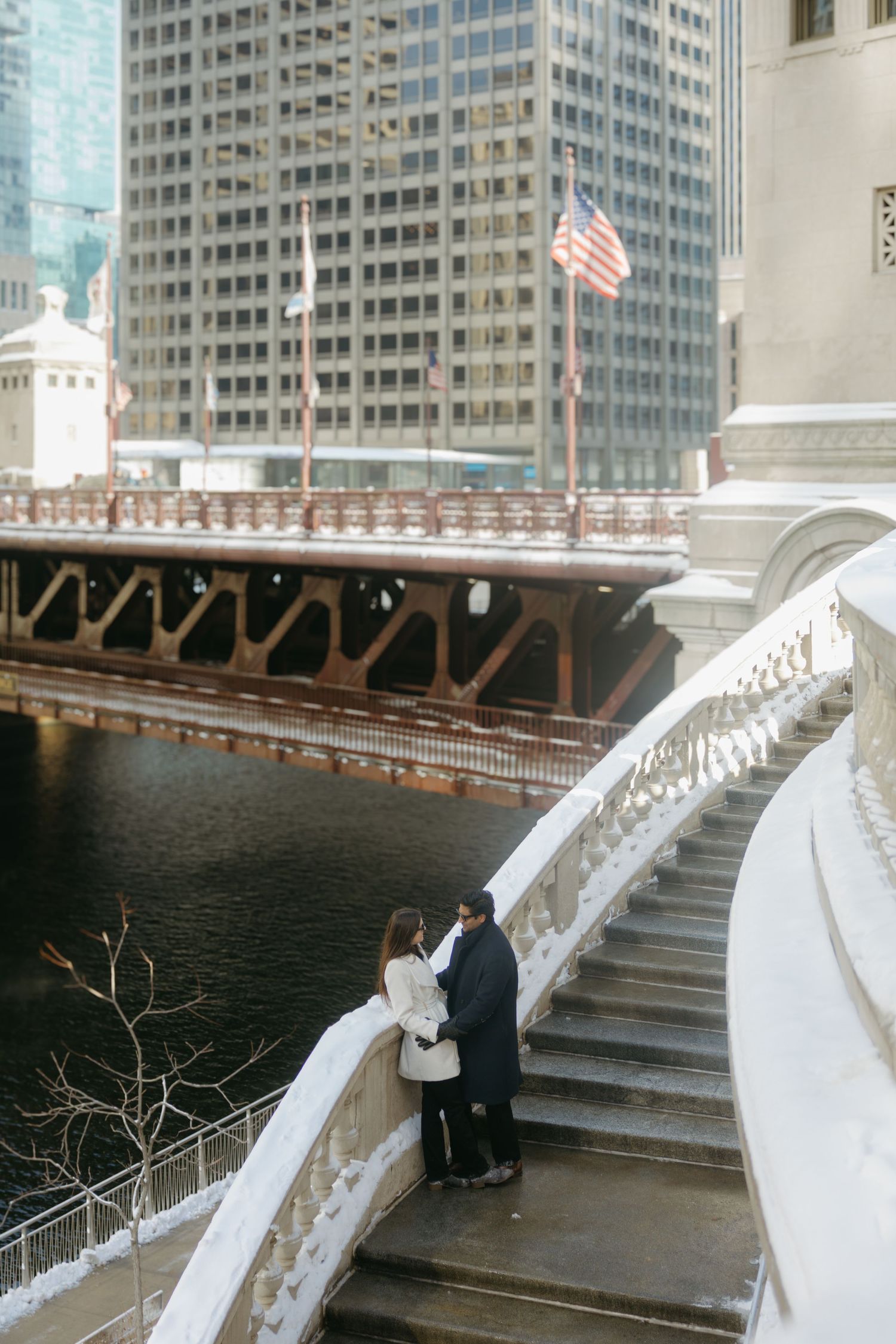 A couple stands on white marble steps overlooking the Chicago River with an American flag and skyscrapers in the background.