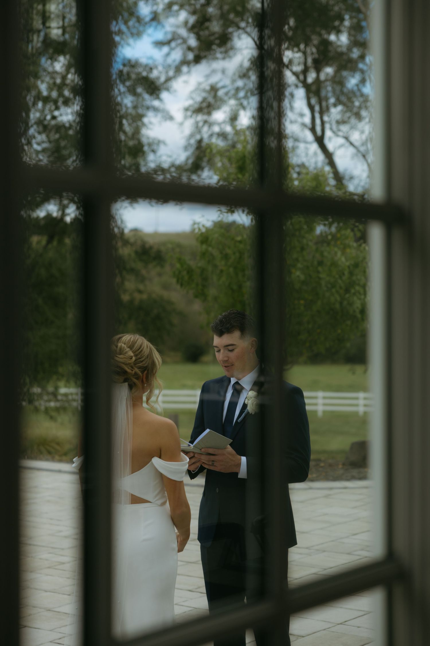 Bride and groom share a tender moment through window panes at an outdoor venue.