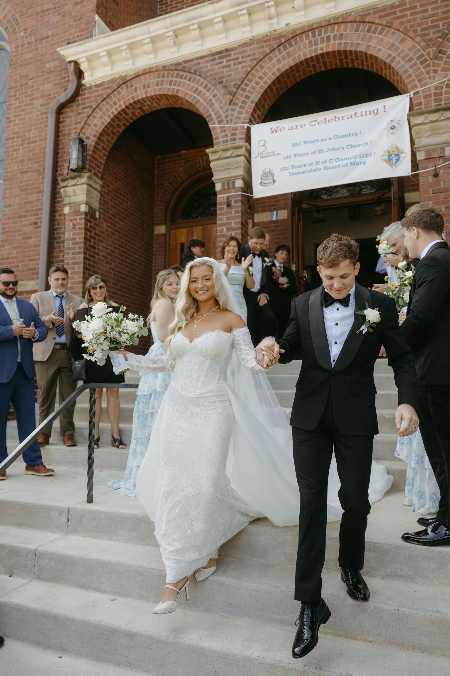 Bride and groom exit brick church holding hands, she wears lace gown with veil, he wears black tuxedo.