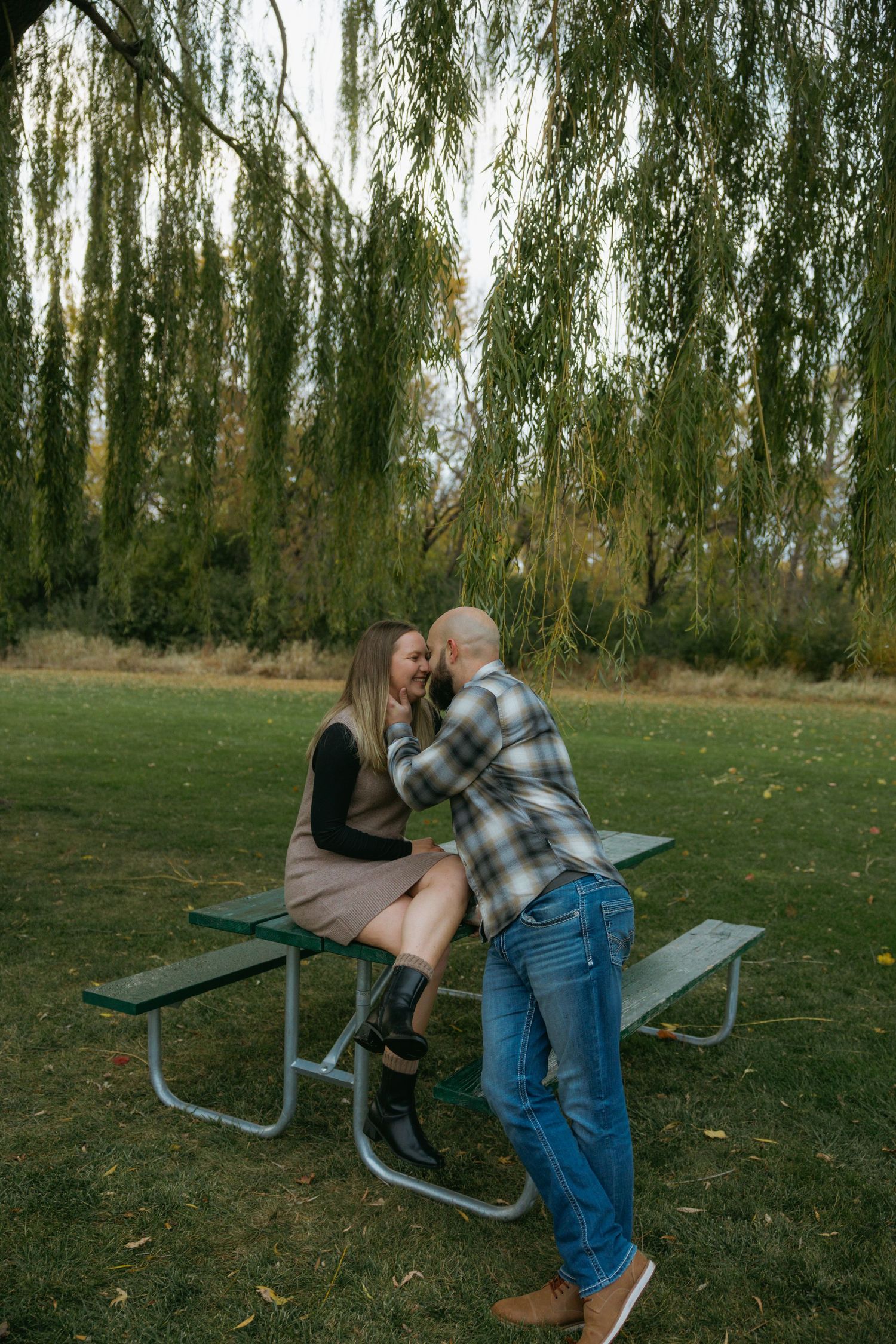 A couple shares a romantic moment on a picnic bench under weeping willow trees in a park setting.