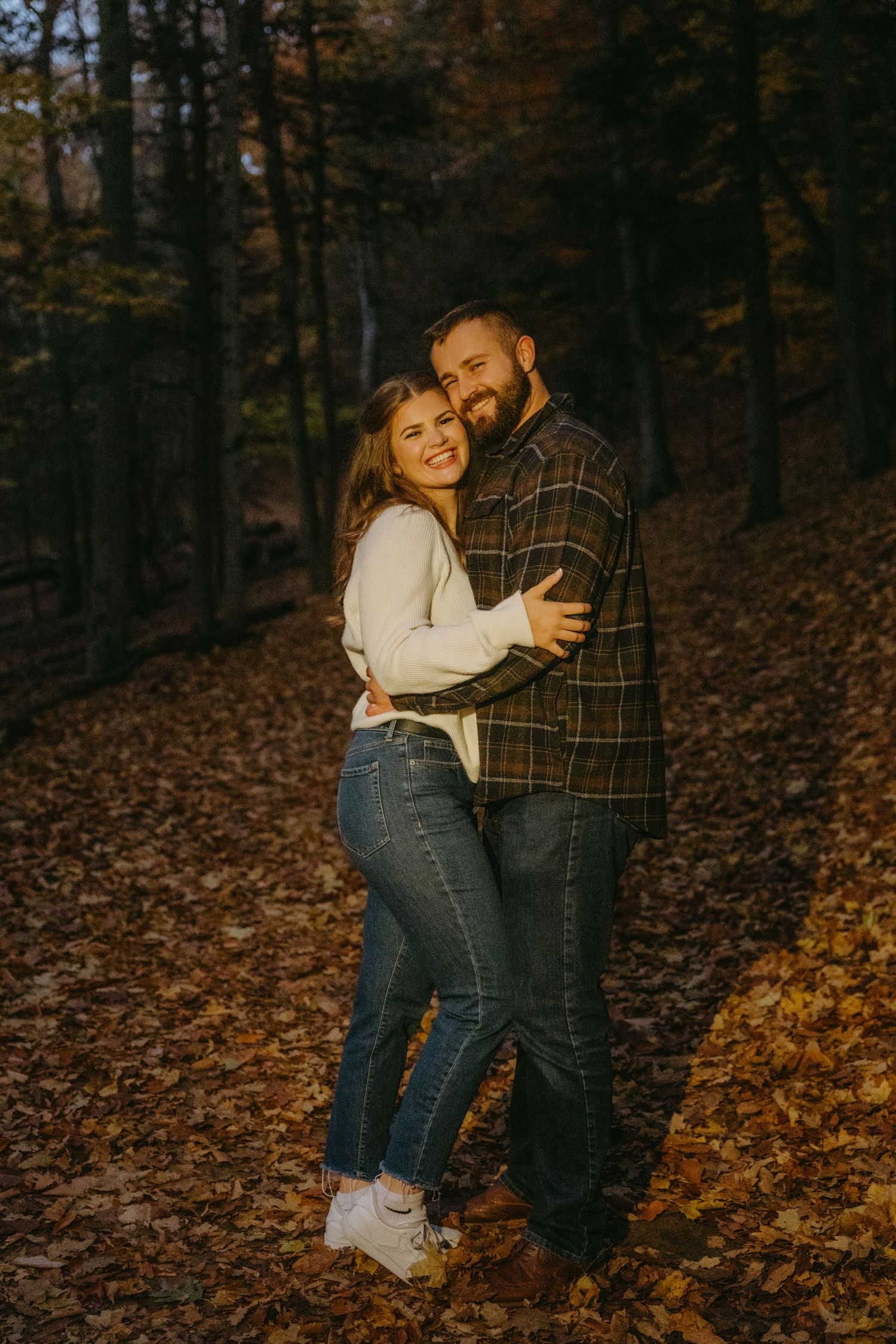 A couple shares a tender moment while sitting together on fallen autumn leaves in a wooded forest setting.