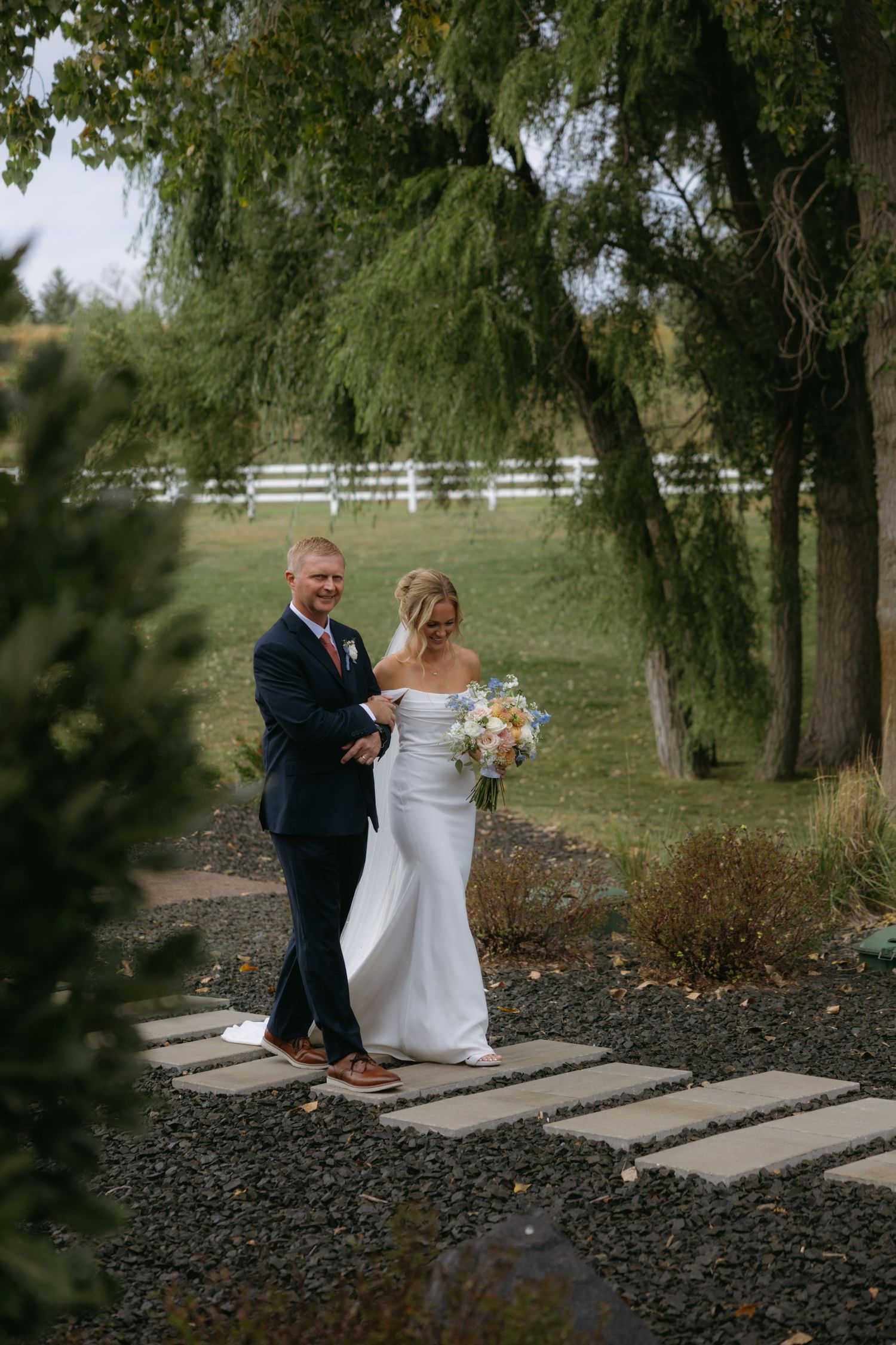 A couple in wedding attire walks together along a stone path surrounded by trees in an outdoor garden setting.