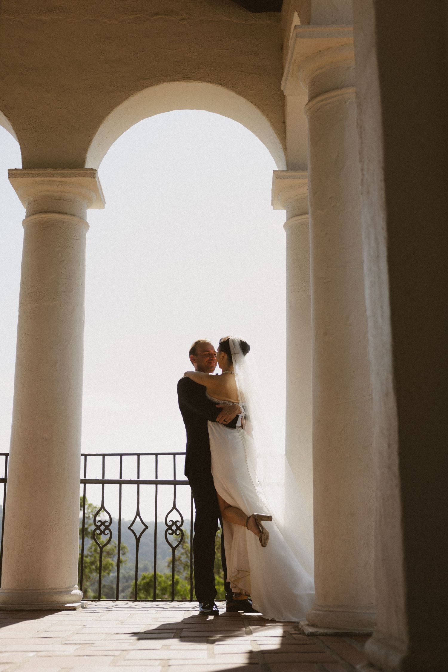 Wedding couple embracing between white arched columns on a sunlit balcony with scenic views.