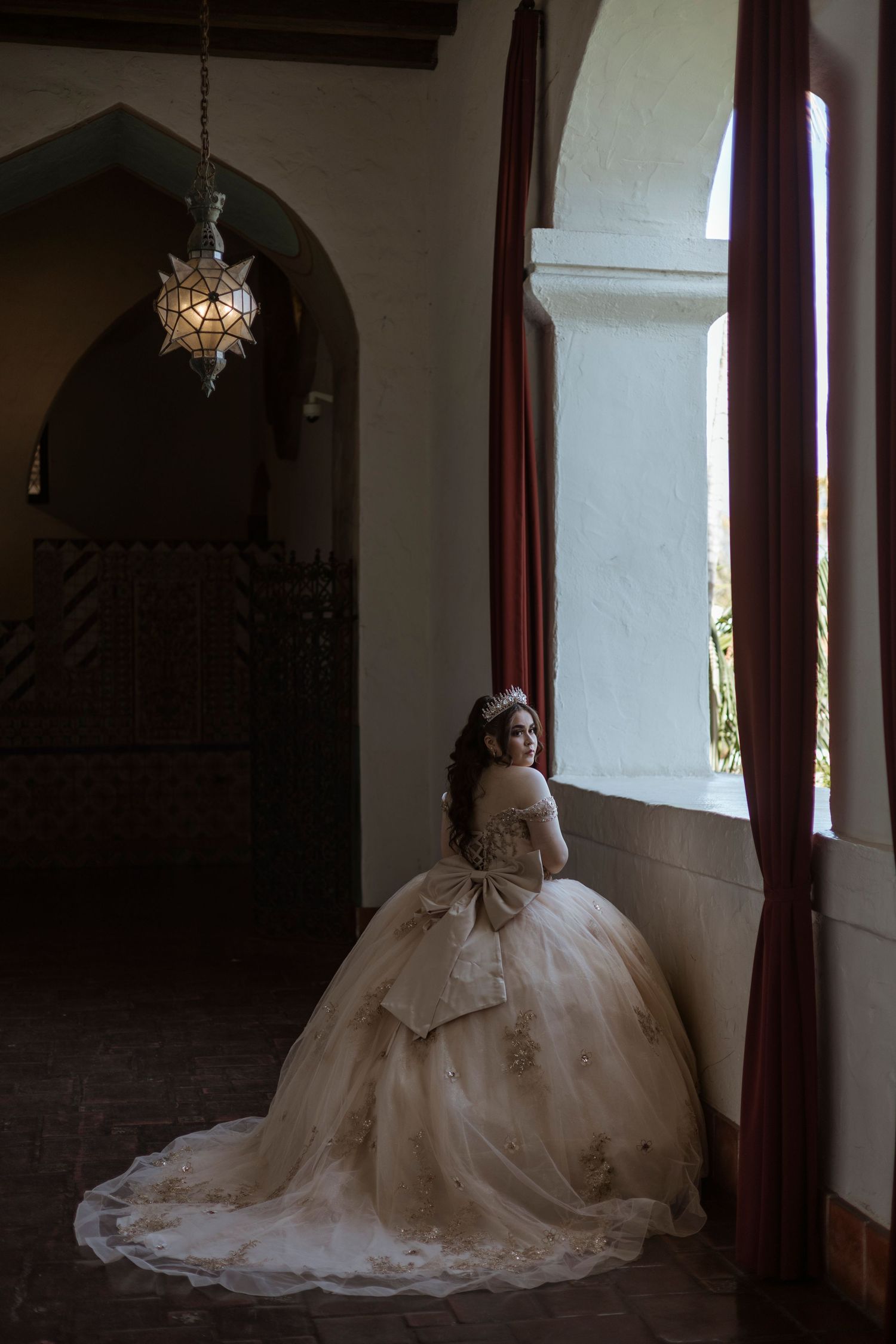 A person in an elegant white wedding gown poses by a window in a gothic archway with dark curtains and vintage decor.