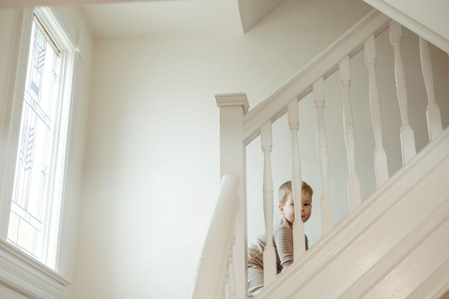 Person peeking through white staircase banister in bright interior with window light creating soft minimalist mood.