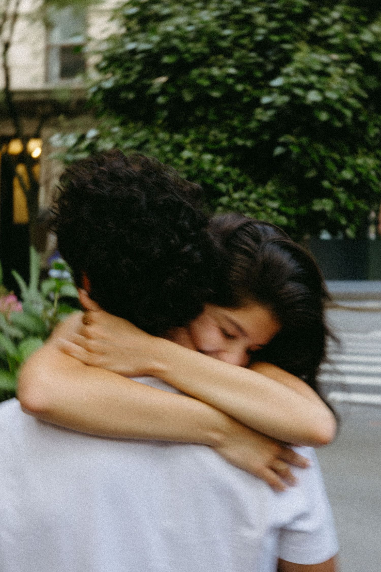 Two people embrace in an intimate hug on a city street with green foliage in the background.