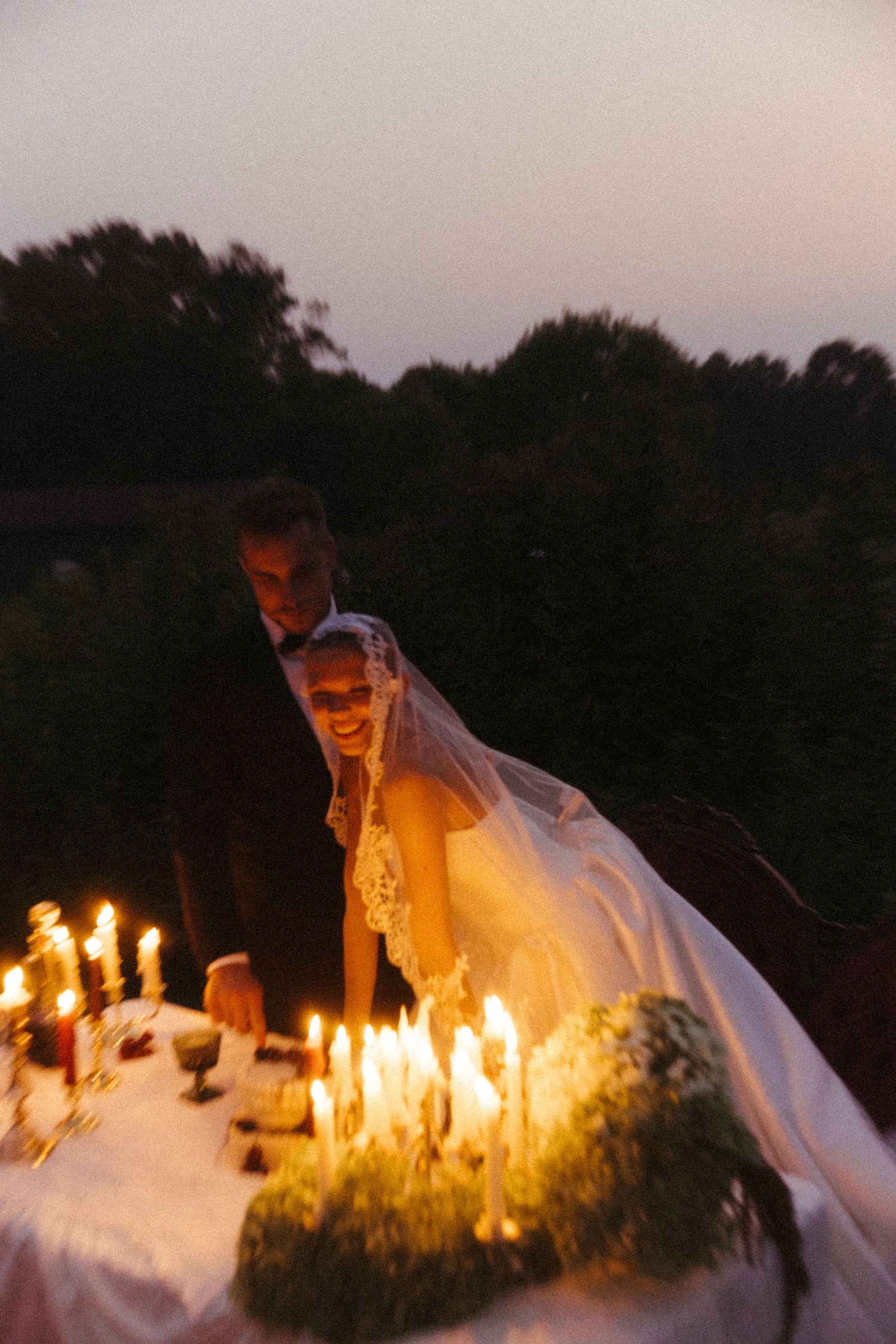 A candlelit outdoor table setting glows warmly at dusk with wedding decorations and flowing white fabric.