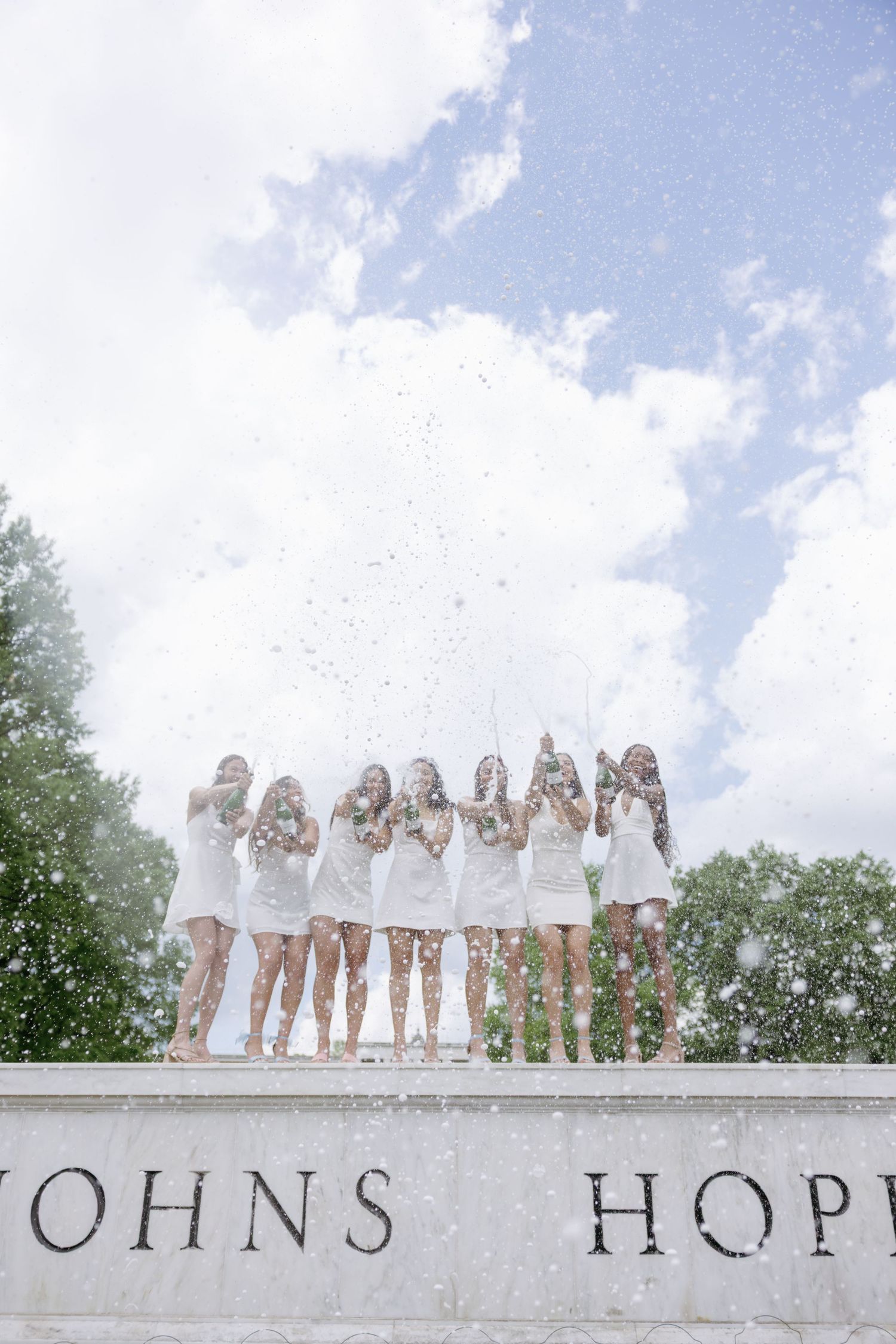 Happy graduates in white dresses stand atop Johns Hopkins monument throwing confetti under blue sky.
