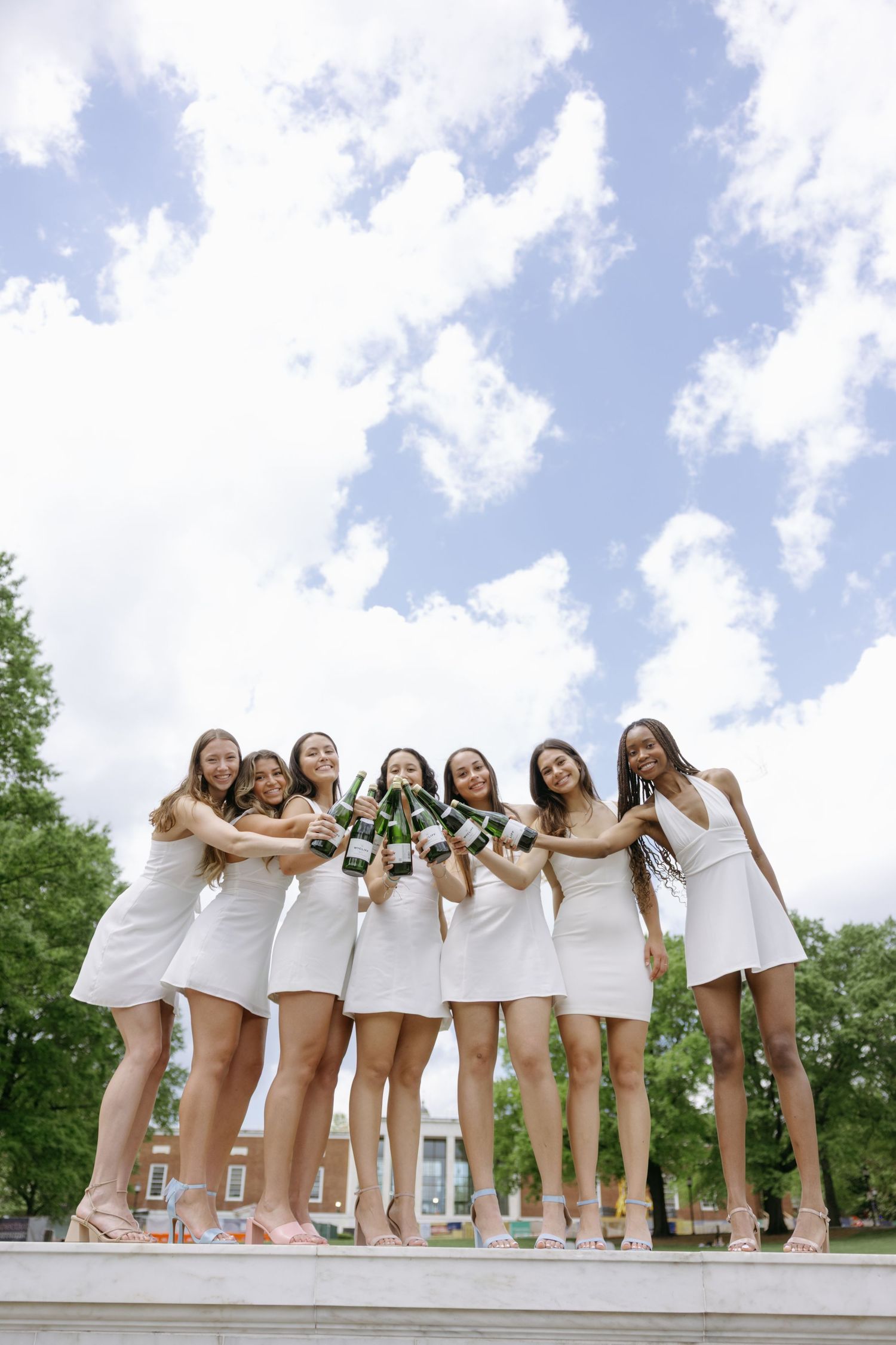 Group in matching white dresses stand together outdoors under blue cloudy sky holding bouquets on a summer day.