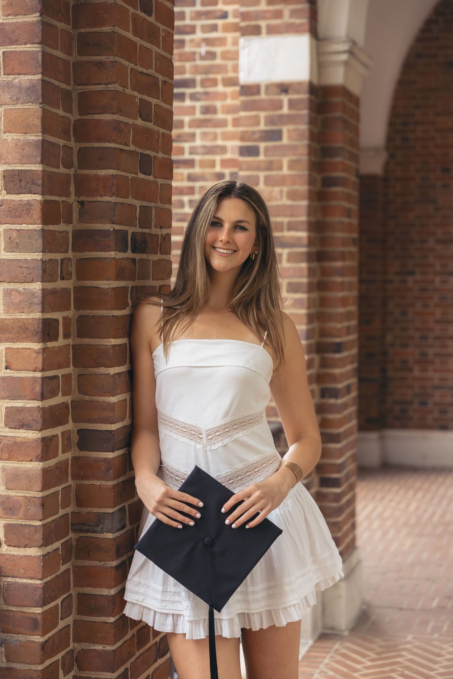 Young woman in white strapless dress holding graduation cap, posing by brick columns at university.