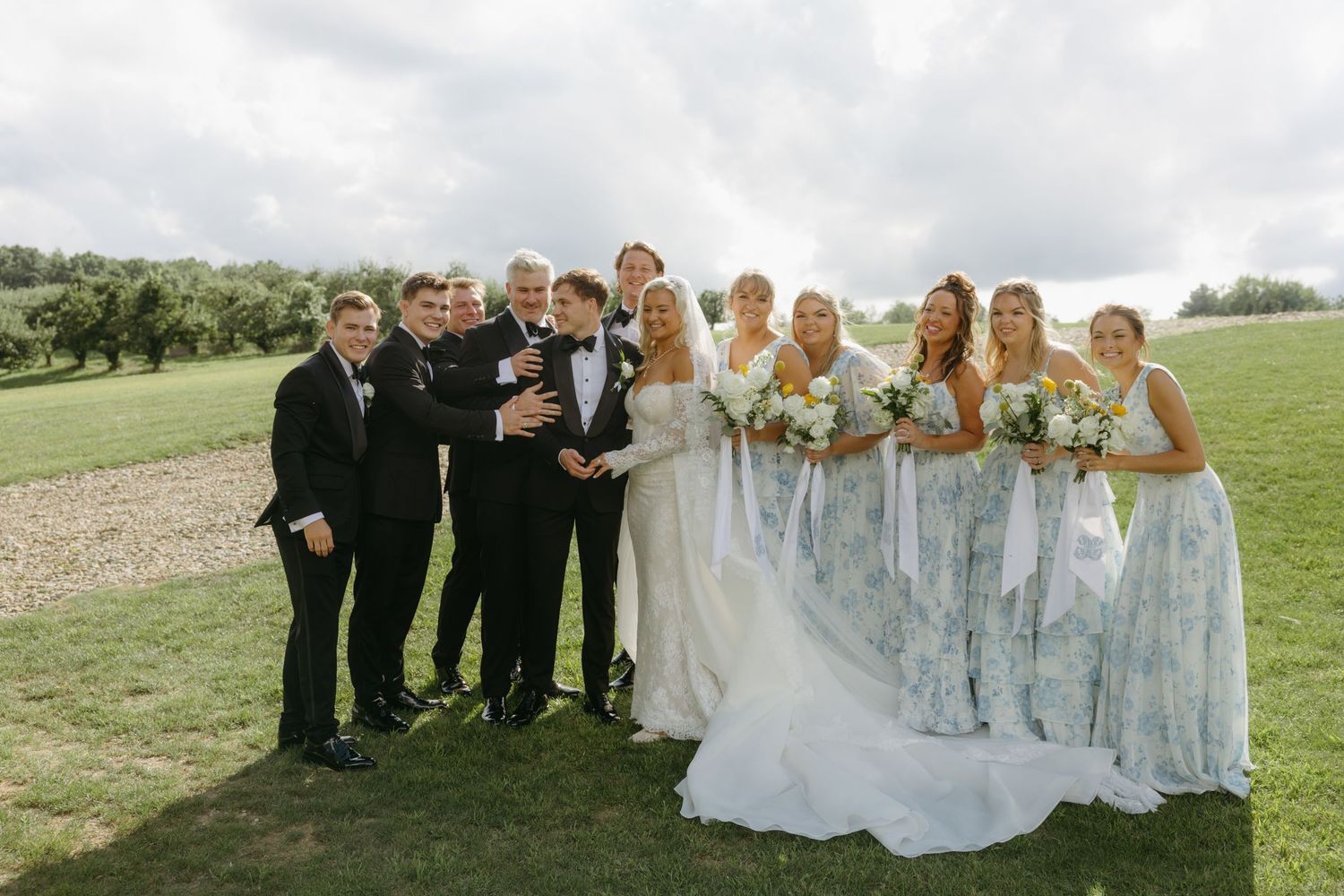 Wedding party poses outdoors on green lawn, bride in white gown with groomsmen in tuxedos and bridesmaids in blue floral dresses.
