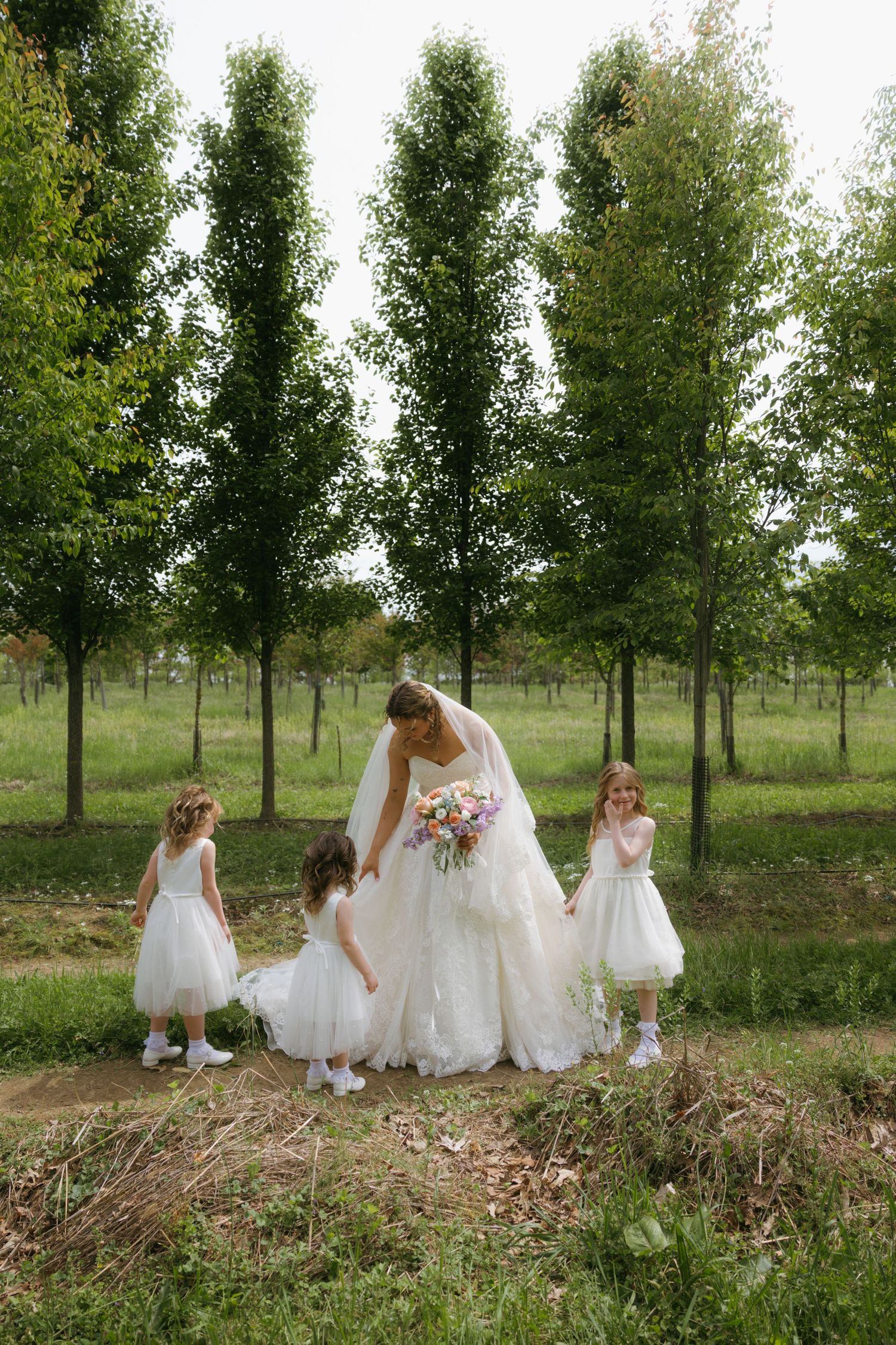Bride in white gown with three flower girls in white dresses standing among tall green trees outdoors.